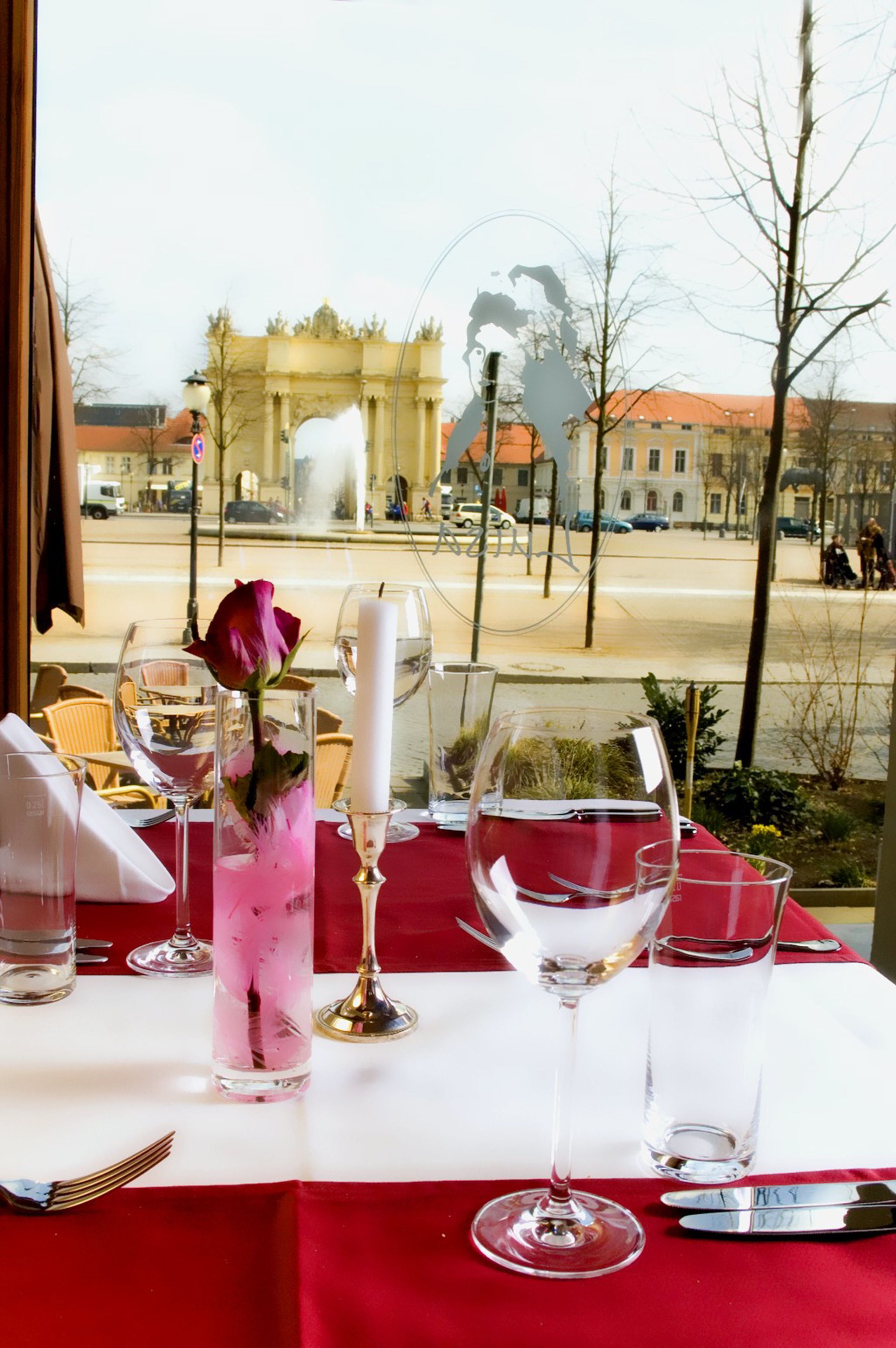 A dining table set with wine glasses, a rose, and a candle, overlooking a street with buildings and a fountain.
