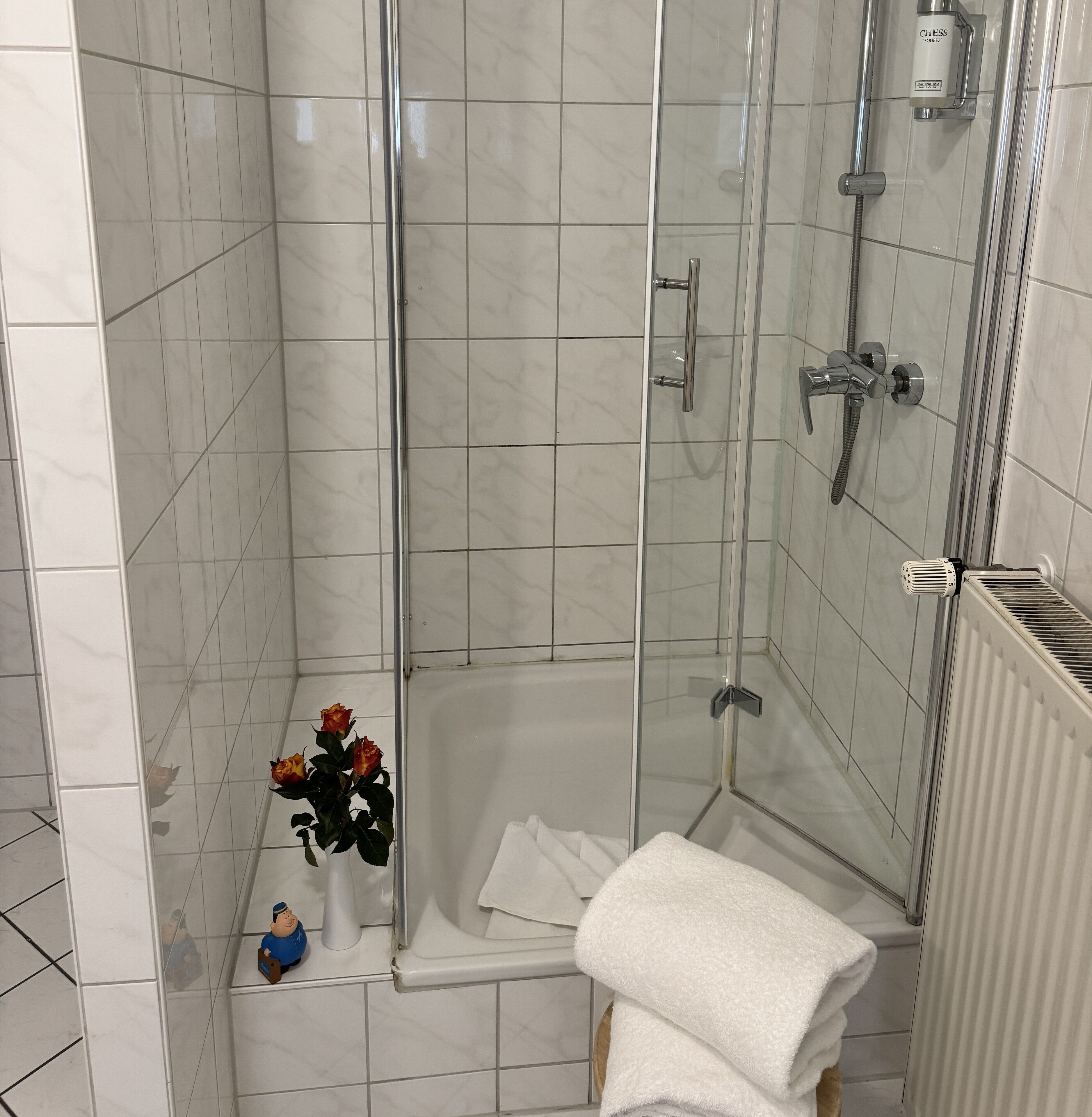 A modern bathroom with a glass-enclosed shower, white tiles, a white bathtub, and a white towel on the floor.