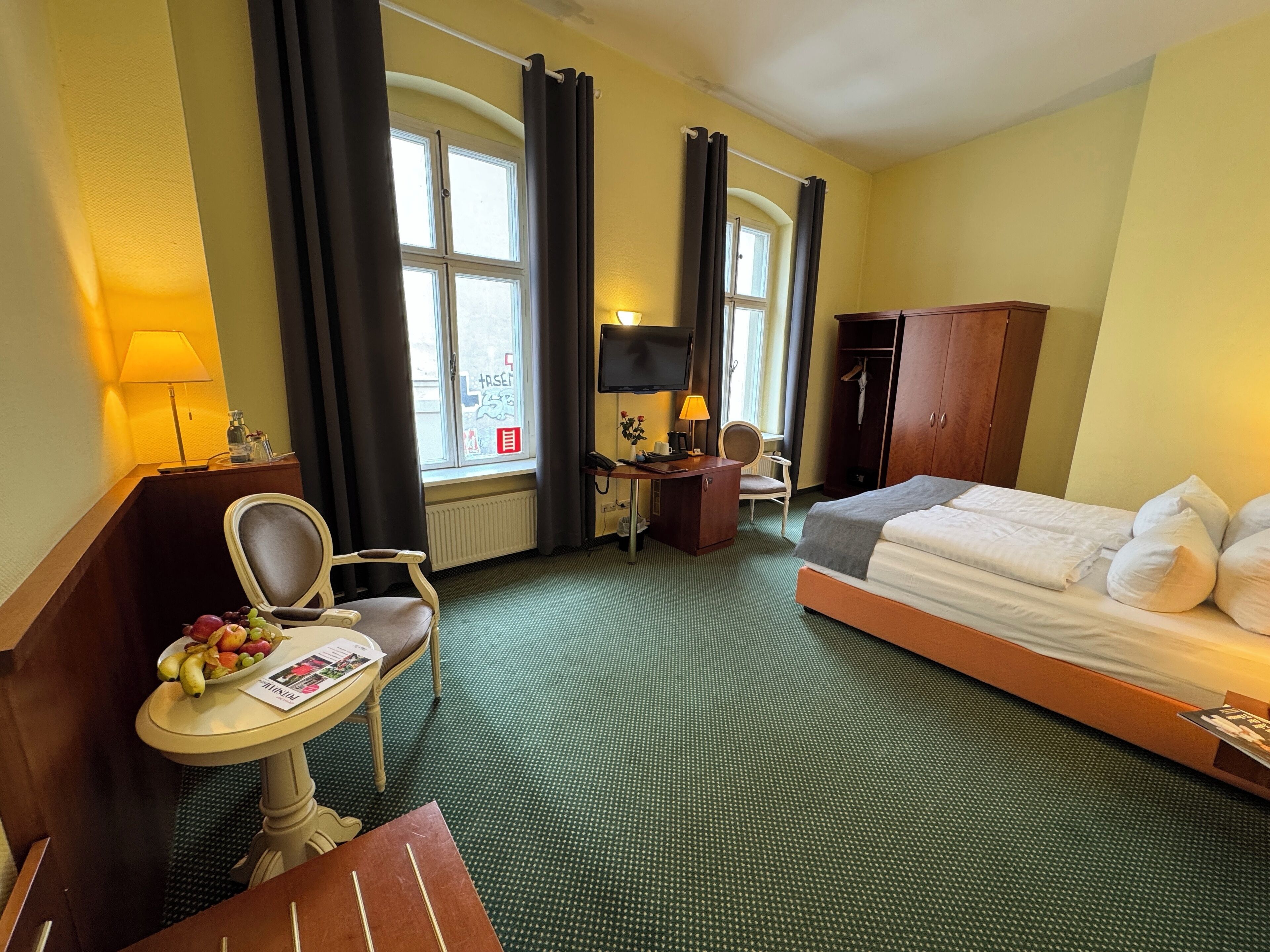 Hotellihuone, jossa on sänky, työpöytä, tuoli, pieni pöytä, jossa on hedelmiä, lamppu ja vaatekaappi.