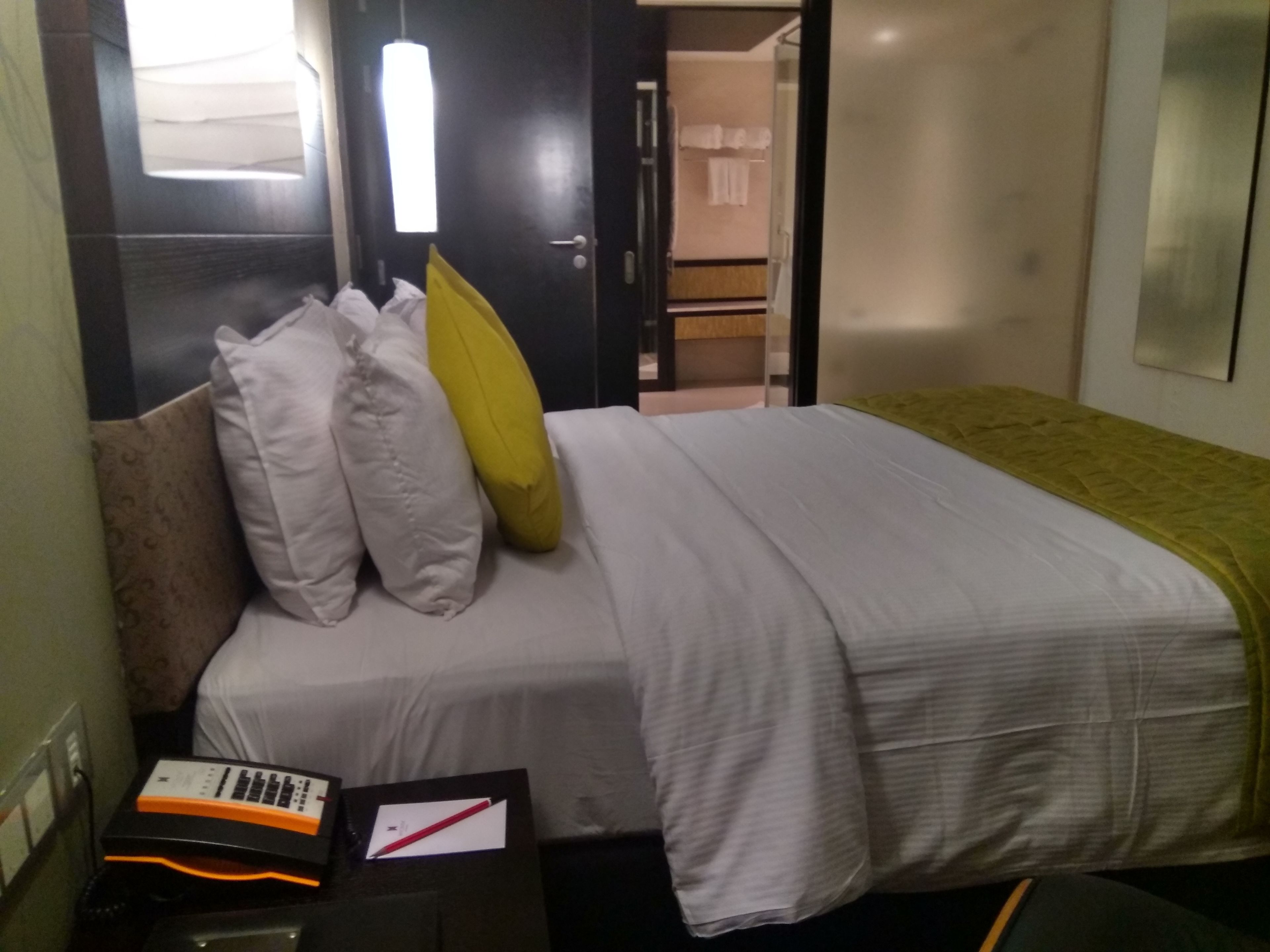 A hotel room with a bed, pillows, a bedside table with a phone and a pen, and a door leading to a bathroom.