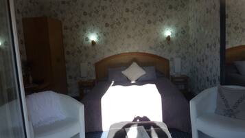 A hotel room with a bed, two bedside tables, a nightstand, a chair, a sofa, and a window with sunlight.