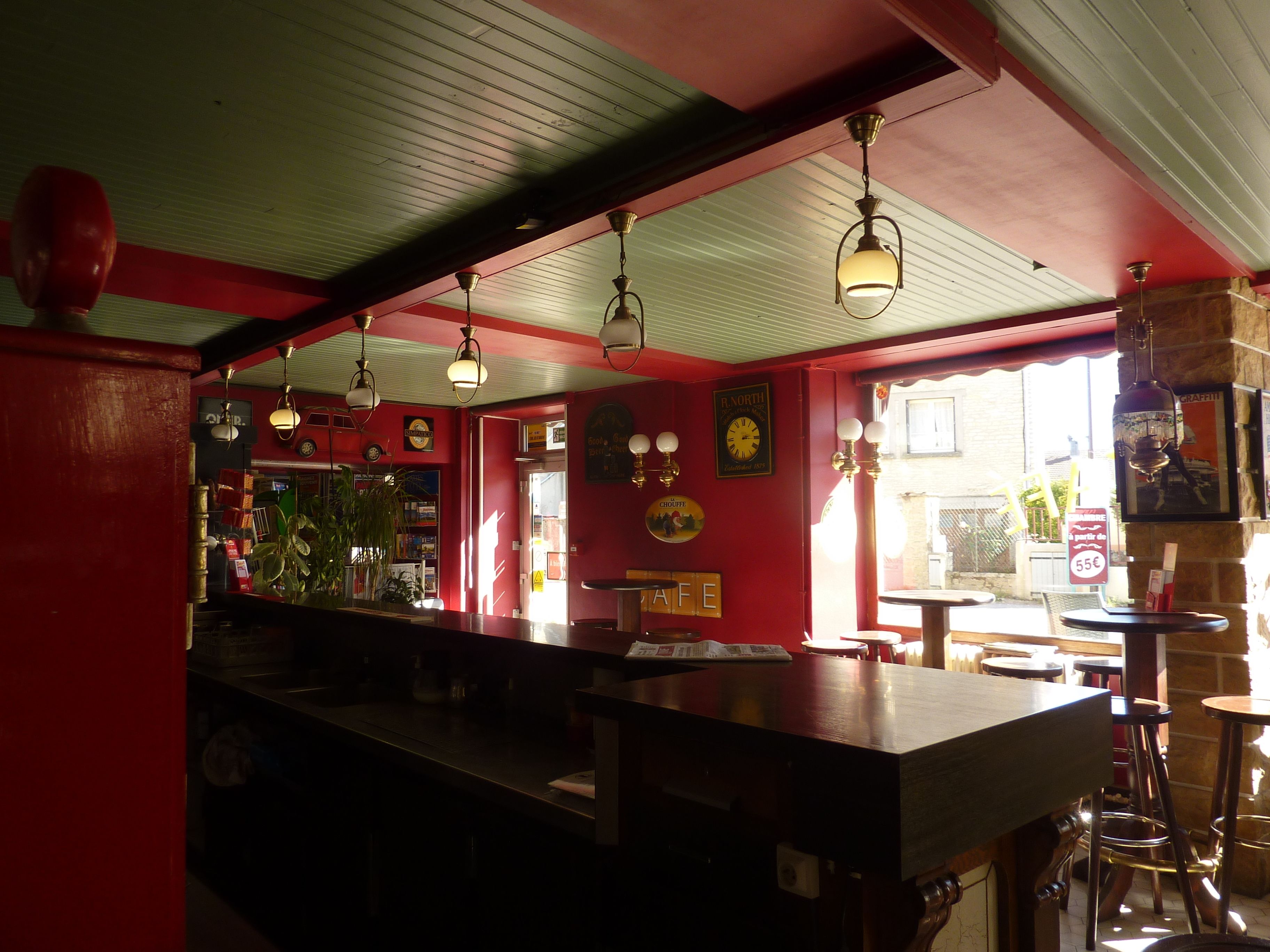 A bar with a red interior, wooden bar counter, hanging lights, and a view of the street outside.
