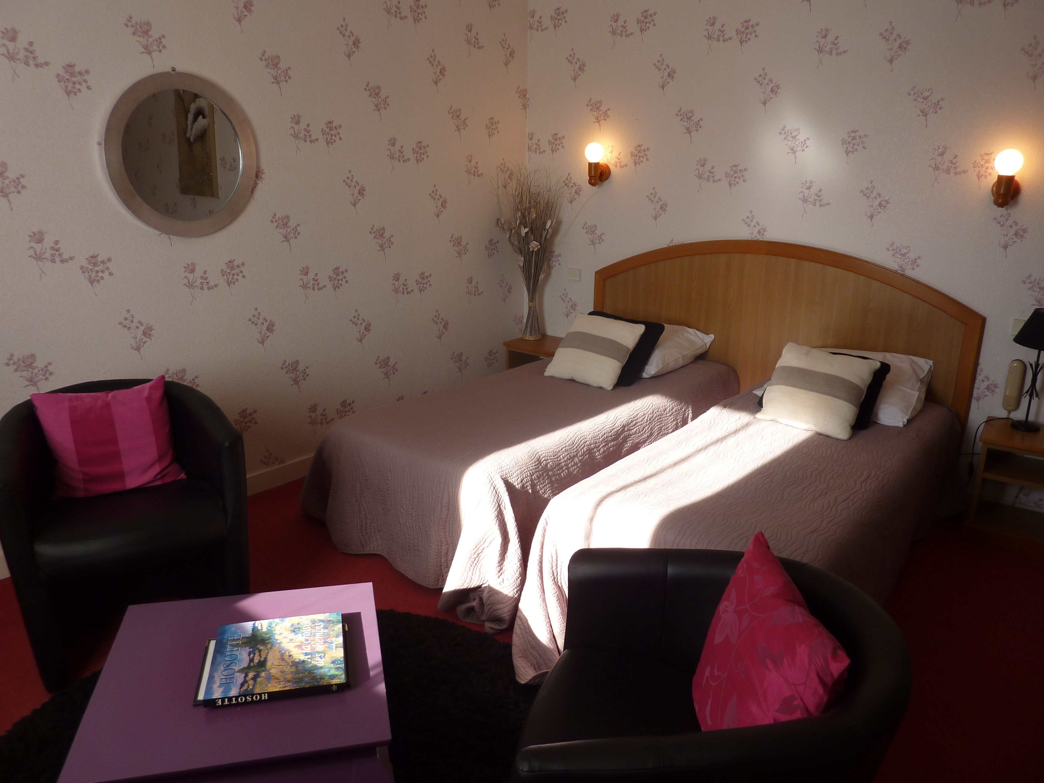 A hotel room with two beds, a round mirror, a small table with a book, and a chair with a pink pillow.