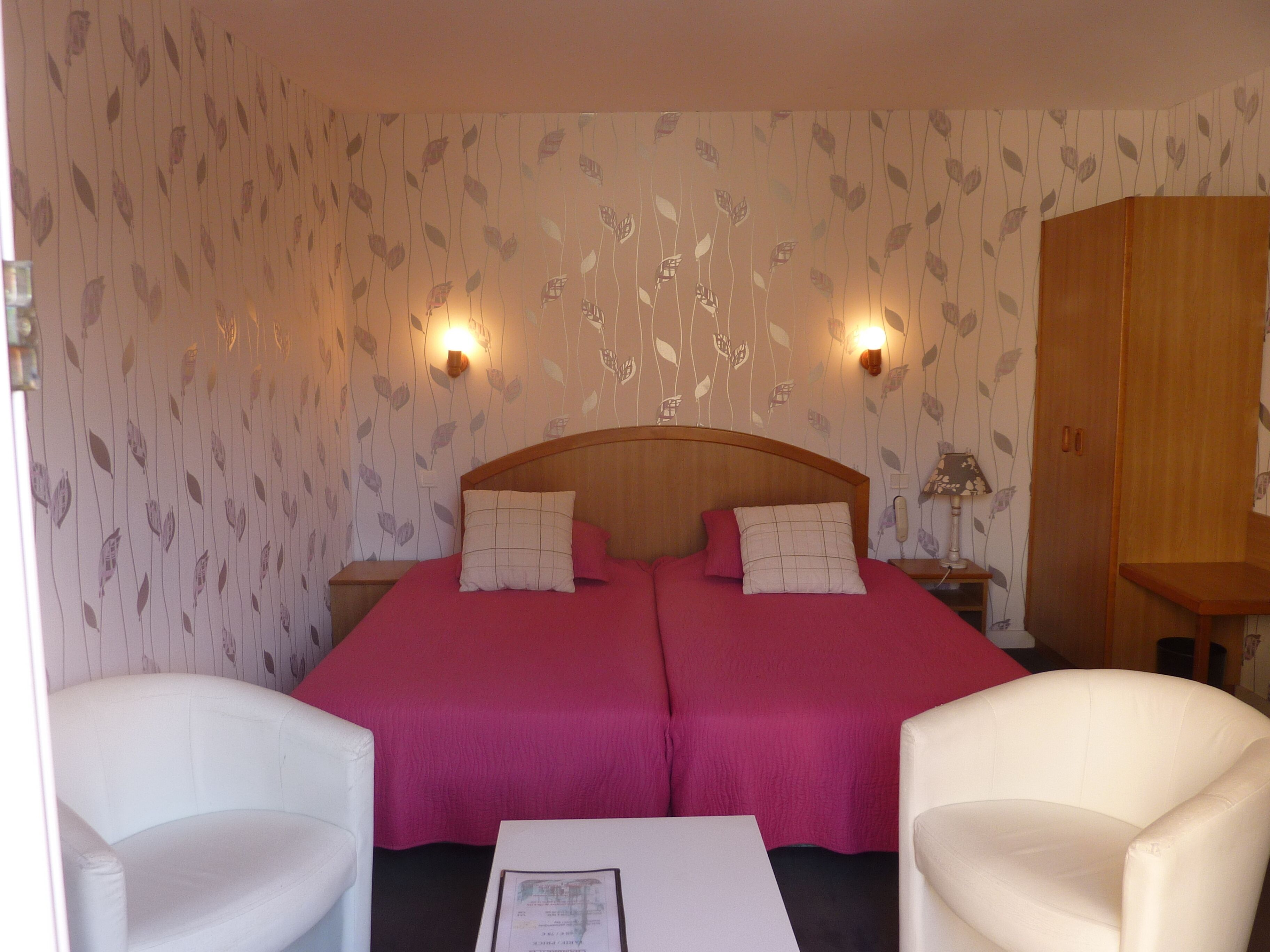 A double bed with two pillows, a nightstand, a lamp, and a wardrobe in a room with leaf-patterned wallpaper.