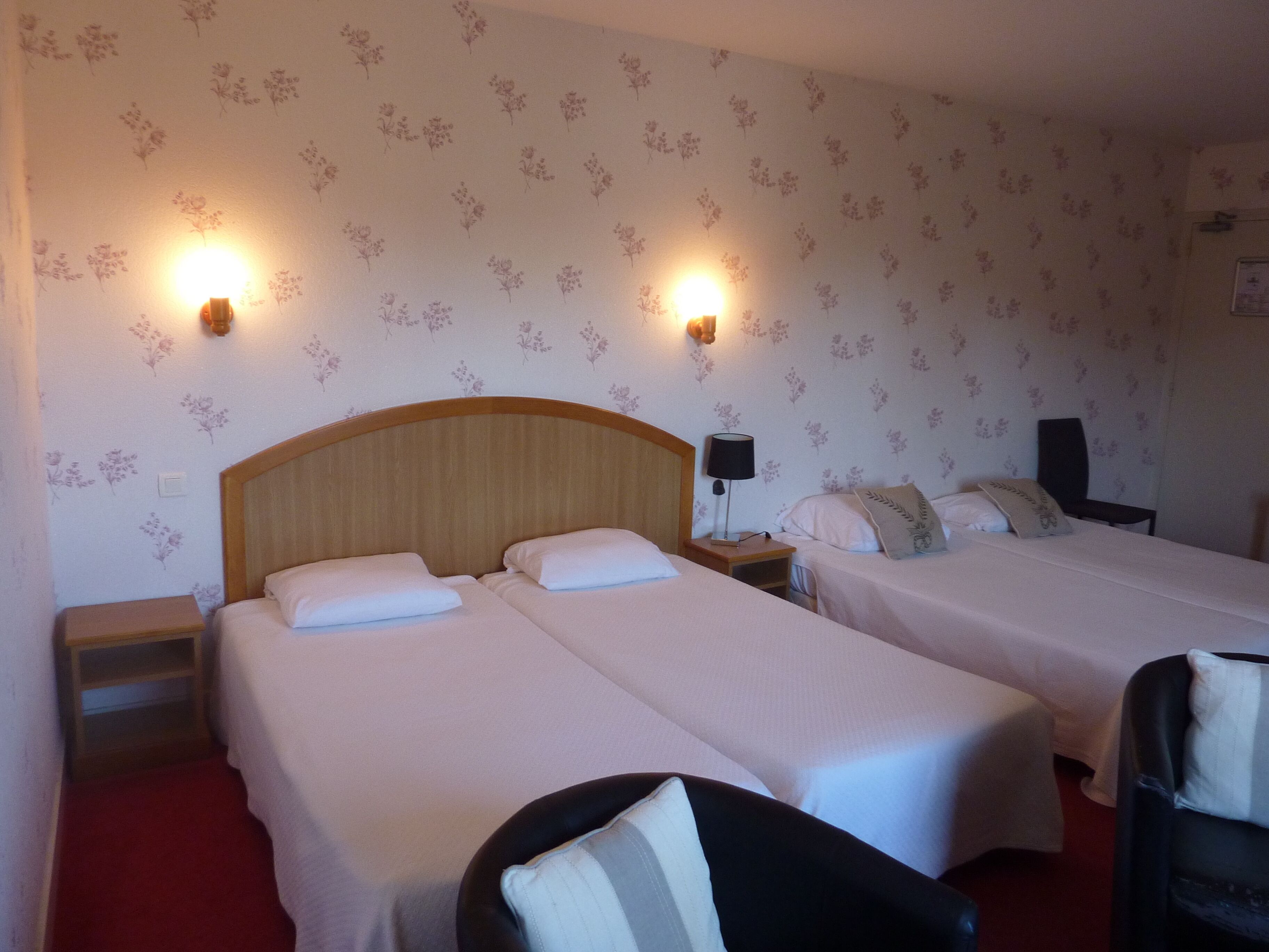 A hotel room with two single beds, a wooden headboard, a nightstand with a lamp, and floral wallpaper.