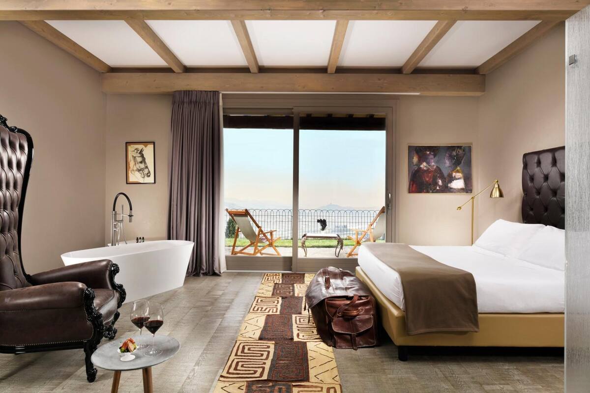 Habitación de hotel con una cama grande, una zona de estar con una silla y una mesa, una bañera y un balcón con vistas.
