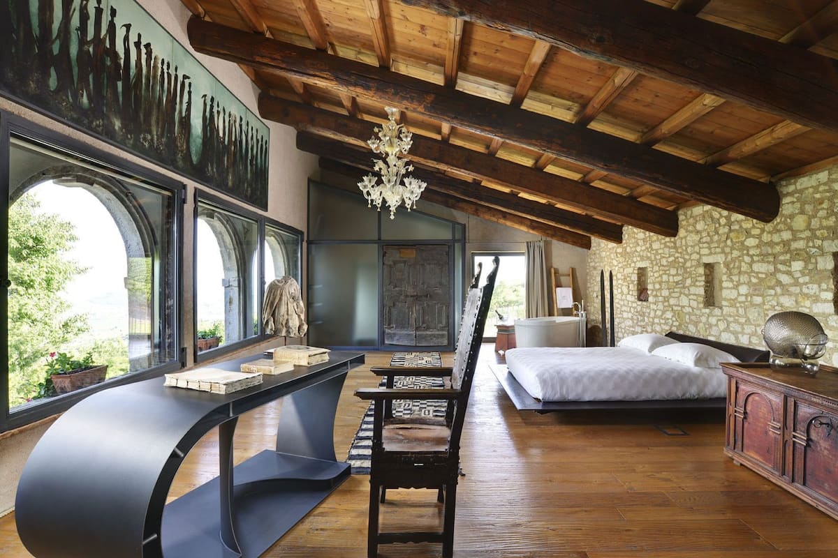 Una habitación con una cama, un escritorio con libros, una araña y una pared de piedra.