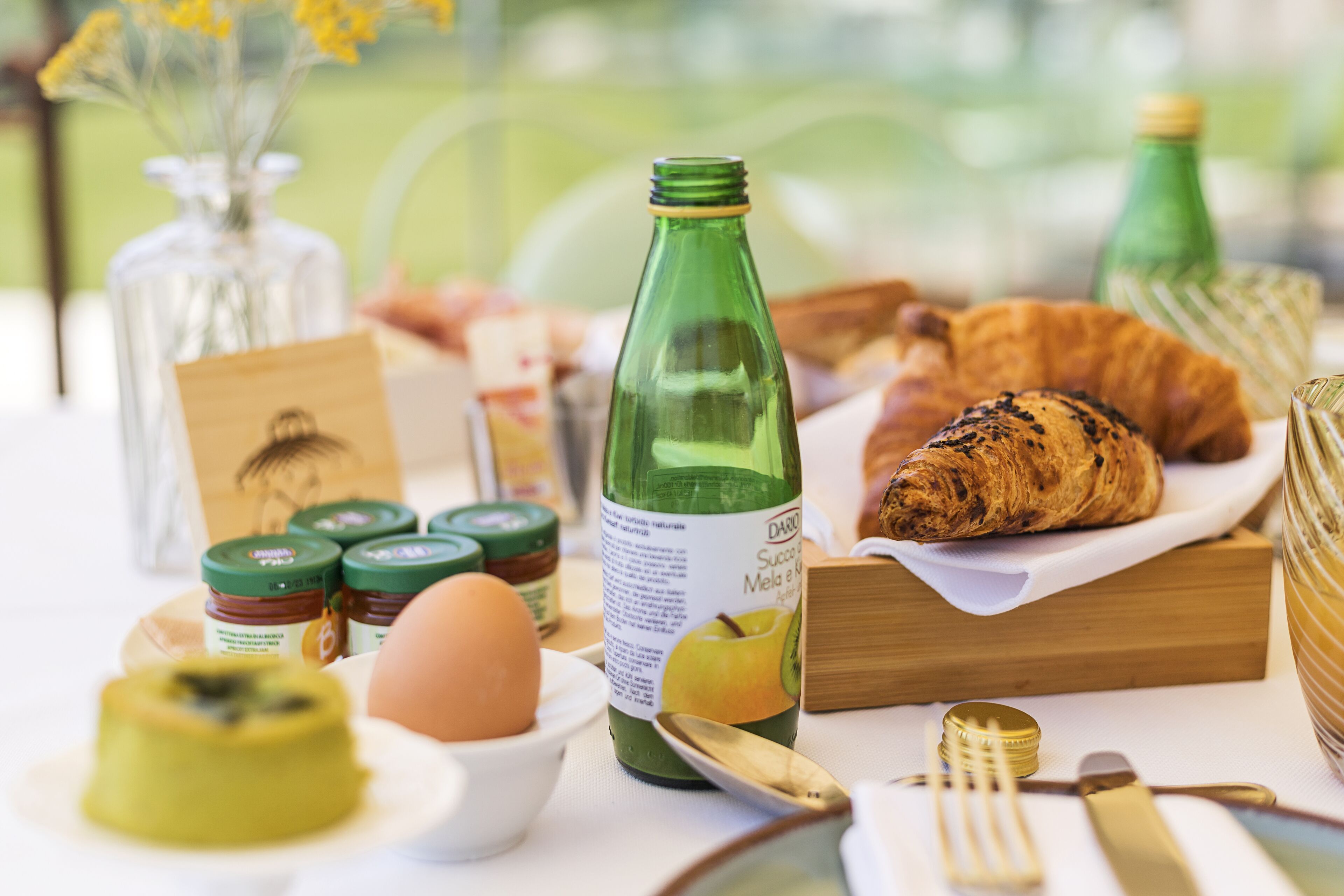 A breakfast table with a bottle, croissant, egg, and jars of jam.