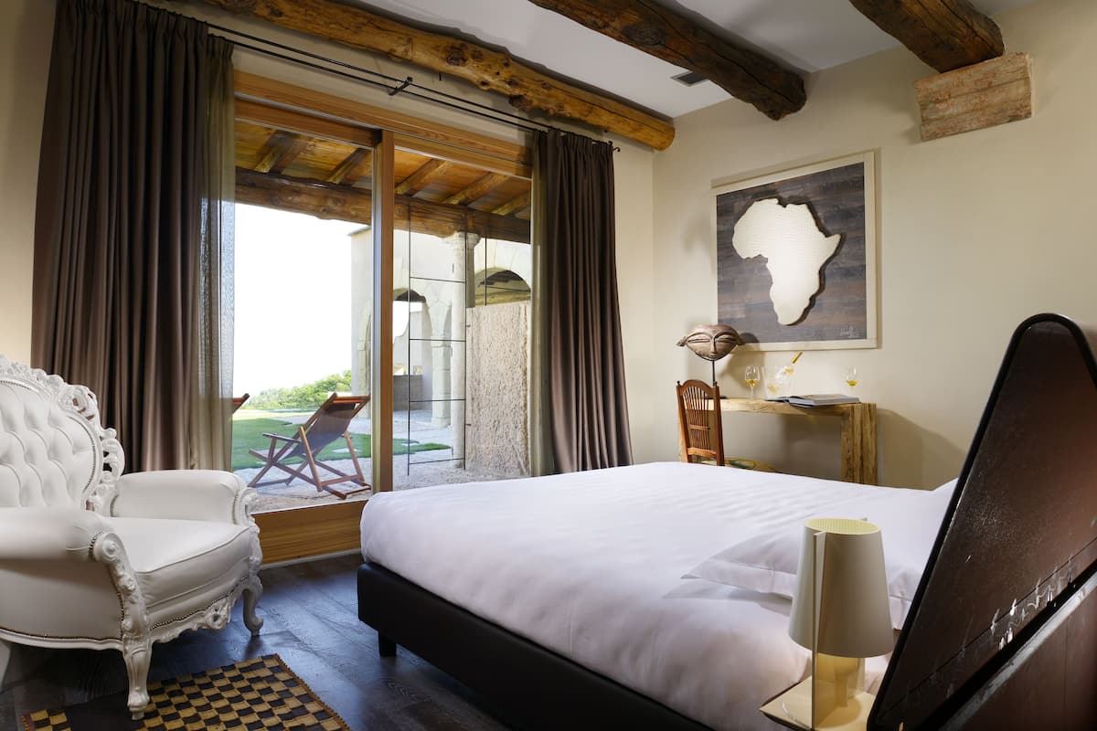 Un dormitorio con una cama grande, un sillón blanco, una mesita de noche de madera con una lámpara, un mapa enmarcado en la pared y un ventanal corredizo que da a un balcón con una silla y una mesa.