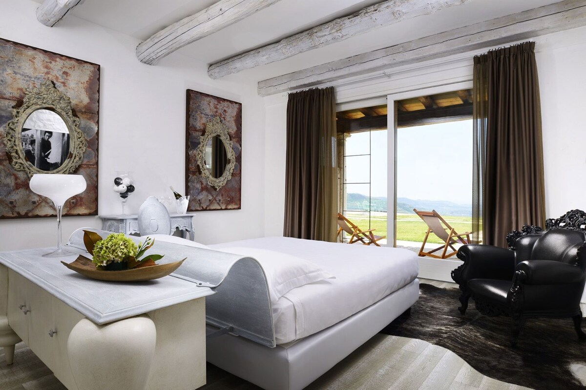 A bedroom with a bed, a desk, a chair, a large window with a view, and artwork on the walls.