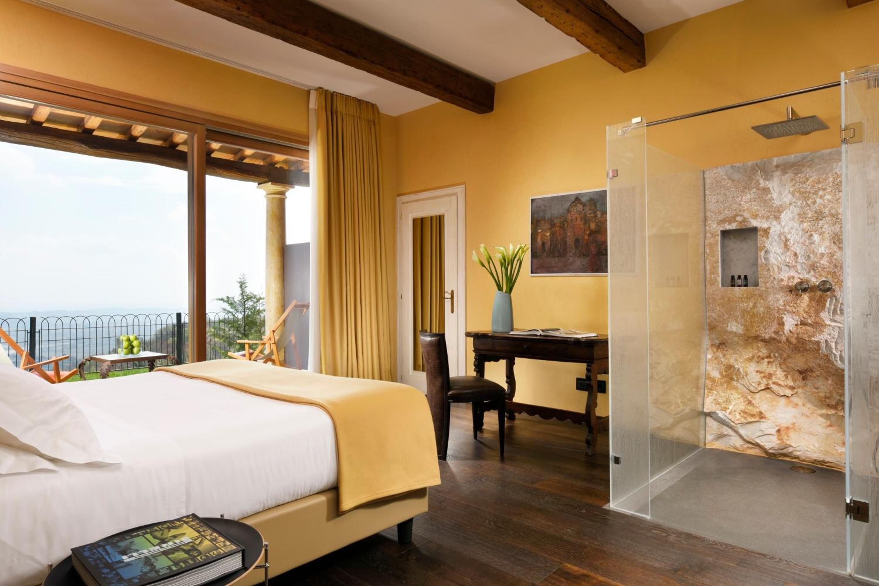 A bedroom with a bed, a desk, a chair, a bookshelf, a painting, and a shower area with a stone wall.