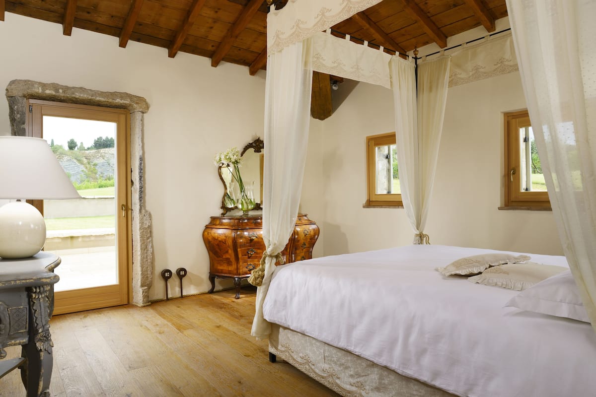 Un dormitorio con techo de madera, una cama con ropa de cama blanca, un aparador de madera y una ventana con cortinas.