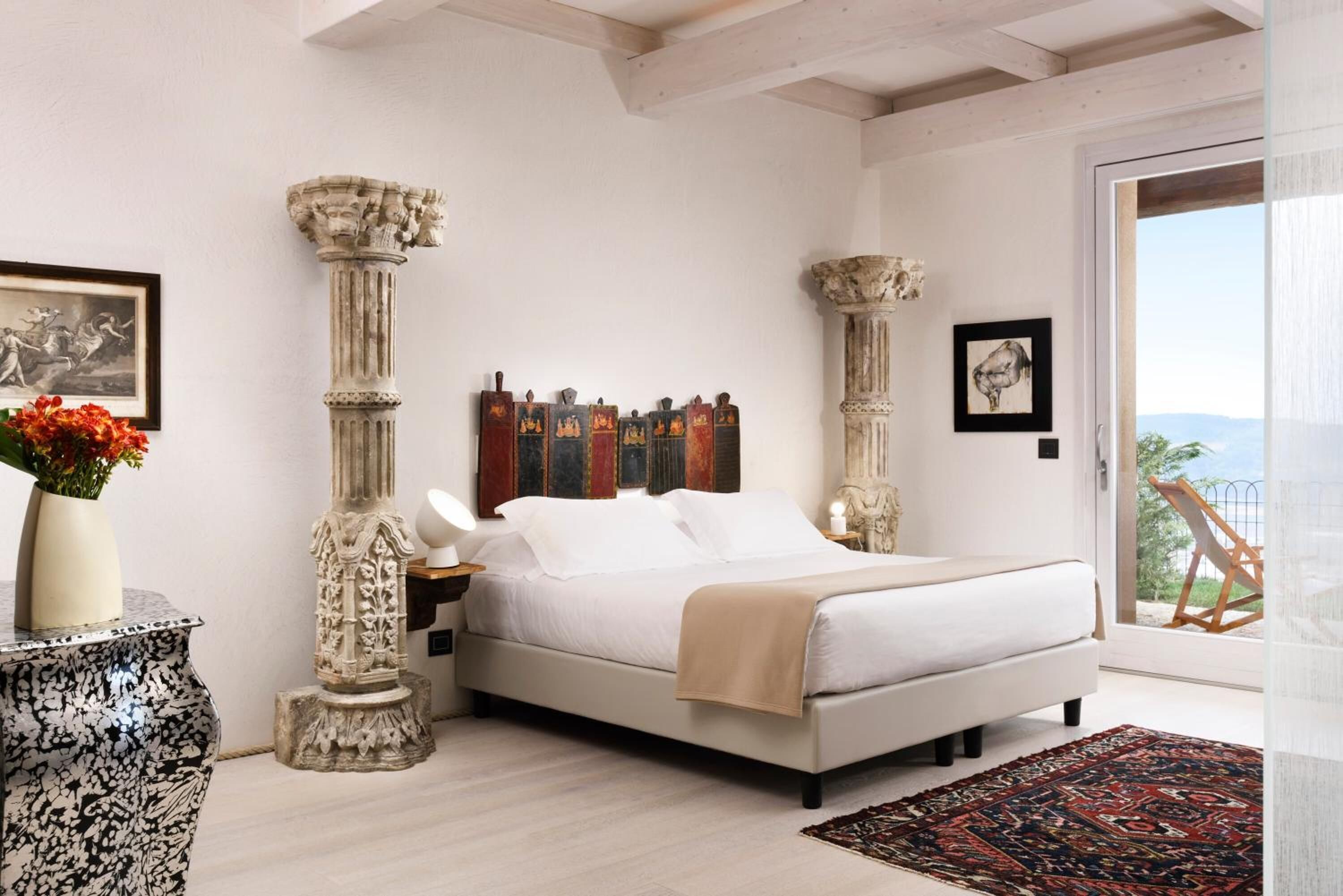 Un dormitorio con cama, mesita de noche, un jarrón con flores y dos columnas.