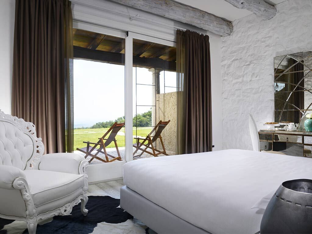 A bedroom with a large bed, a white armchair, a desk, and a view of the outdoors through a sliding door.