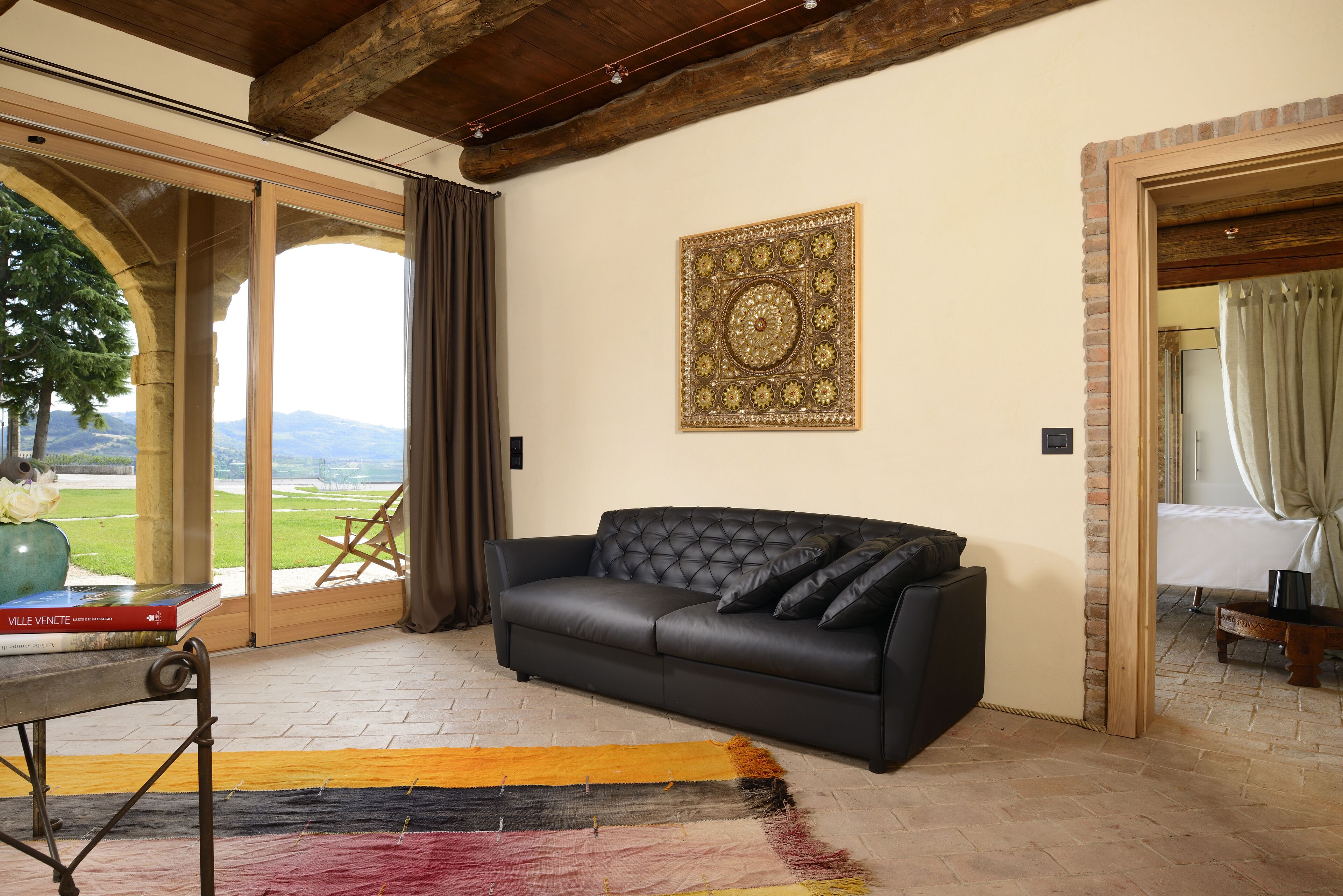 A room with a black leather sofa, a large decorative wall piece, and a view of a green landscape through a glass door.