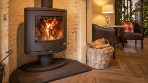 A modern wood-burning stove in a cozy room with a brick wall, a wicker basket of firewood, a table with a lamp, and a leather chair. - Bankton House Hotel (Livingston)