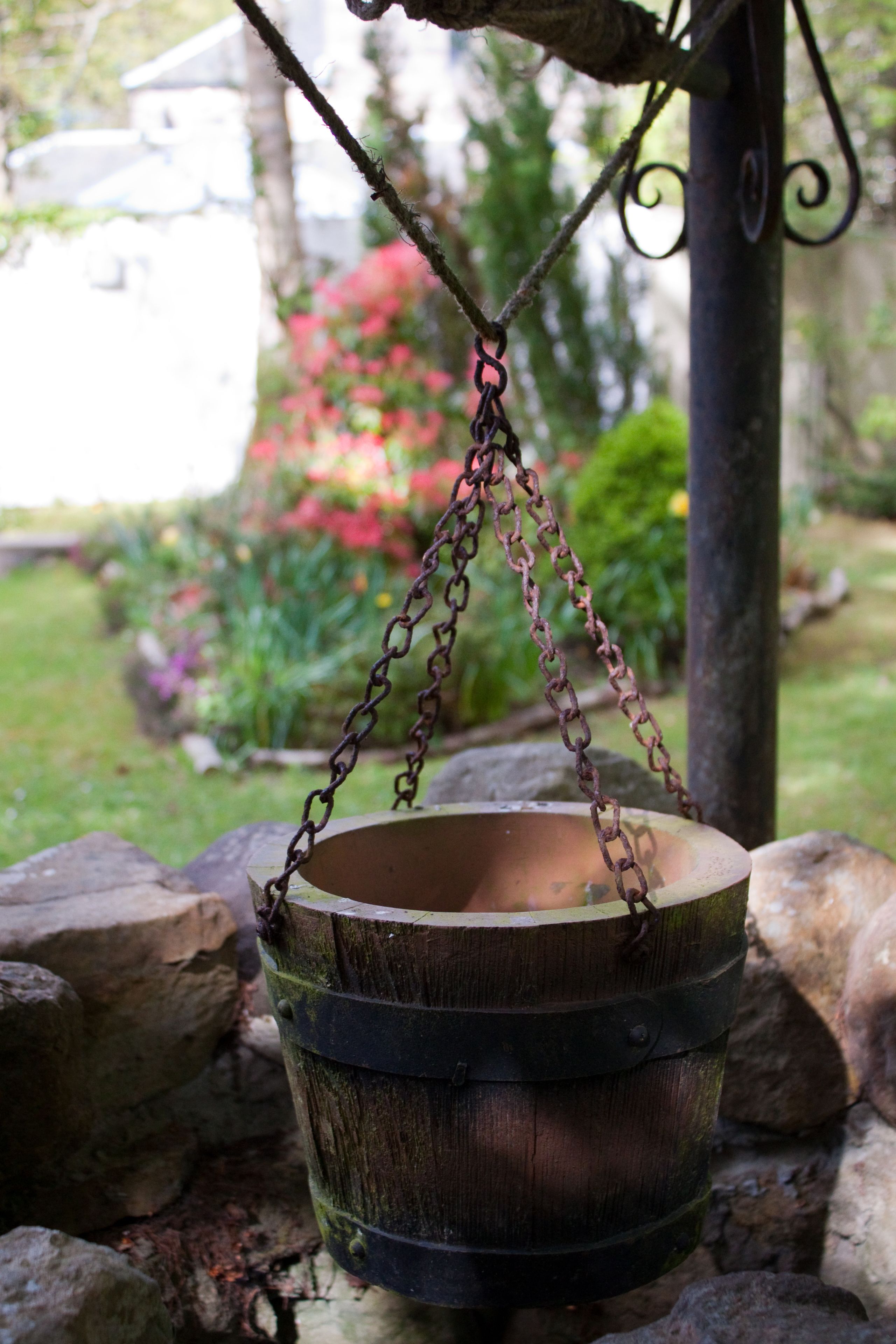 A rustic metal bucket hanging from a chain, surrounded by rocks and greenery.