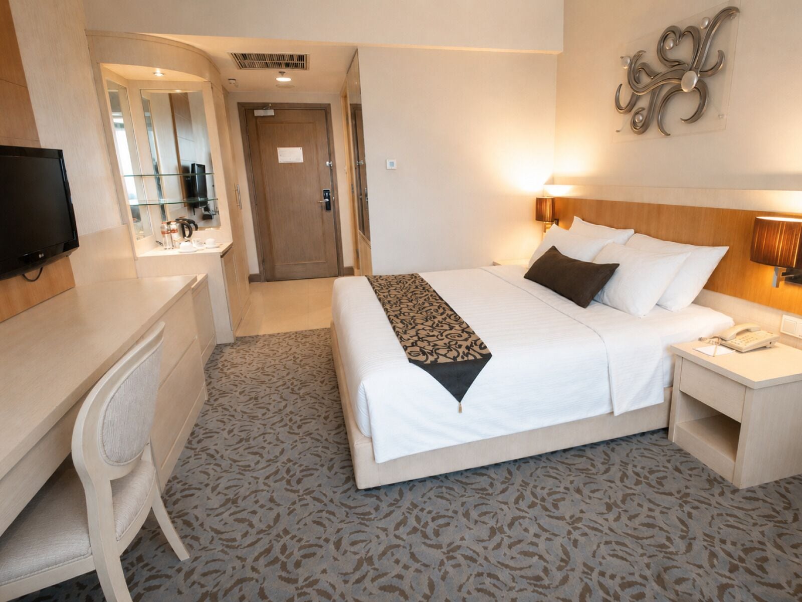A hotel room with a bed, bedside table, lamp, and a desk with a chair.