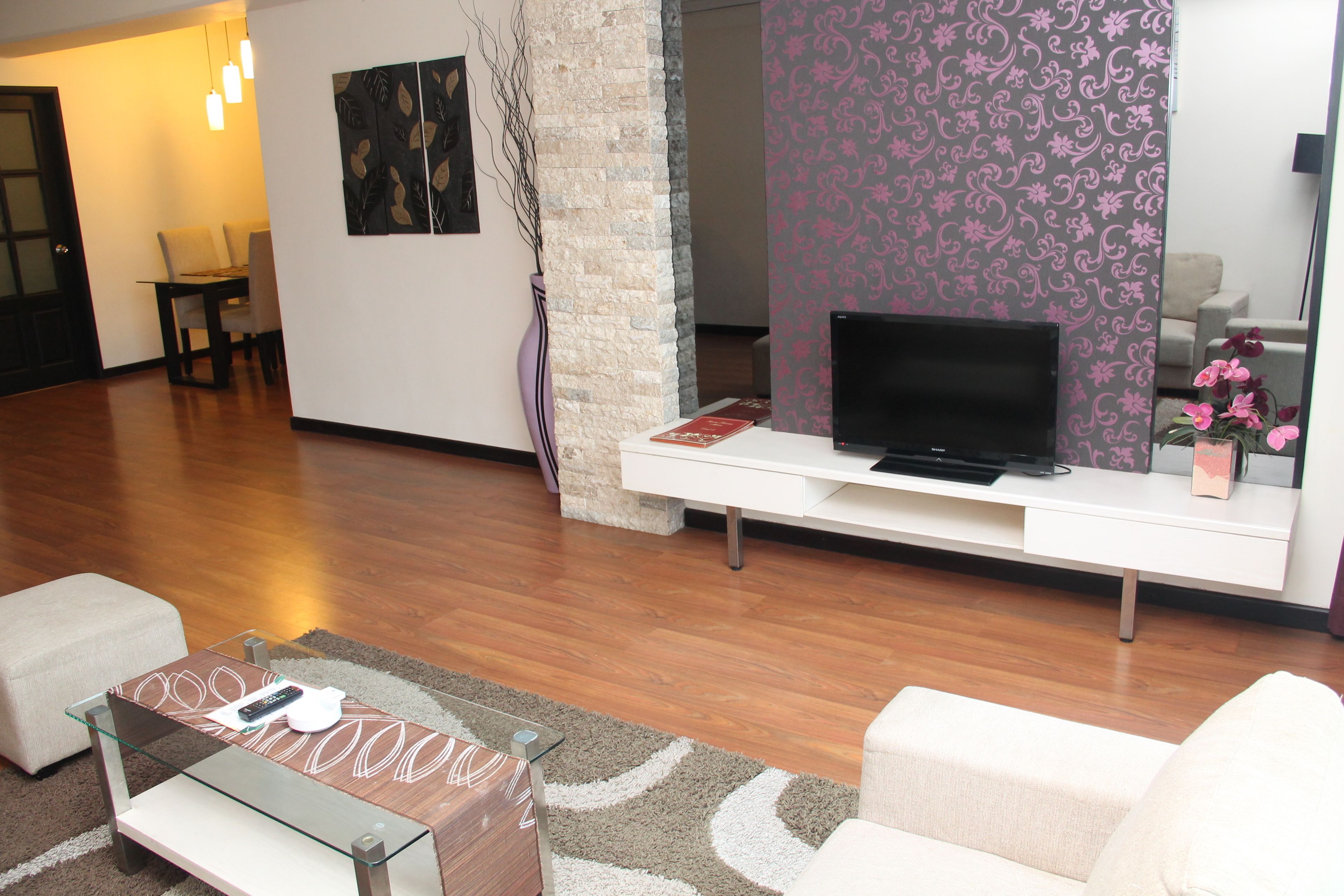 Un salon moderne avec un téléviseur à écran plat posé sur une console blanche, une table basse en verre et un canapé beige.