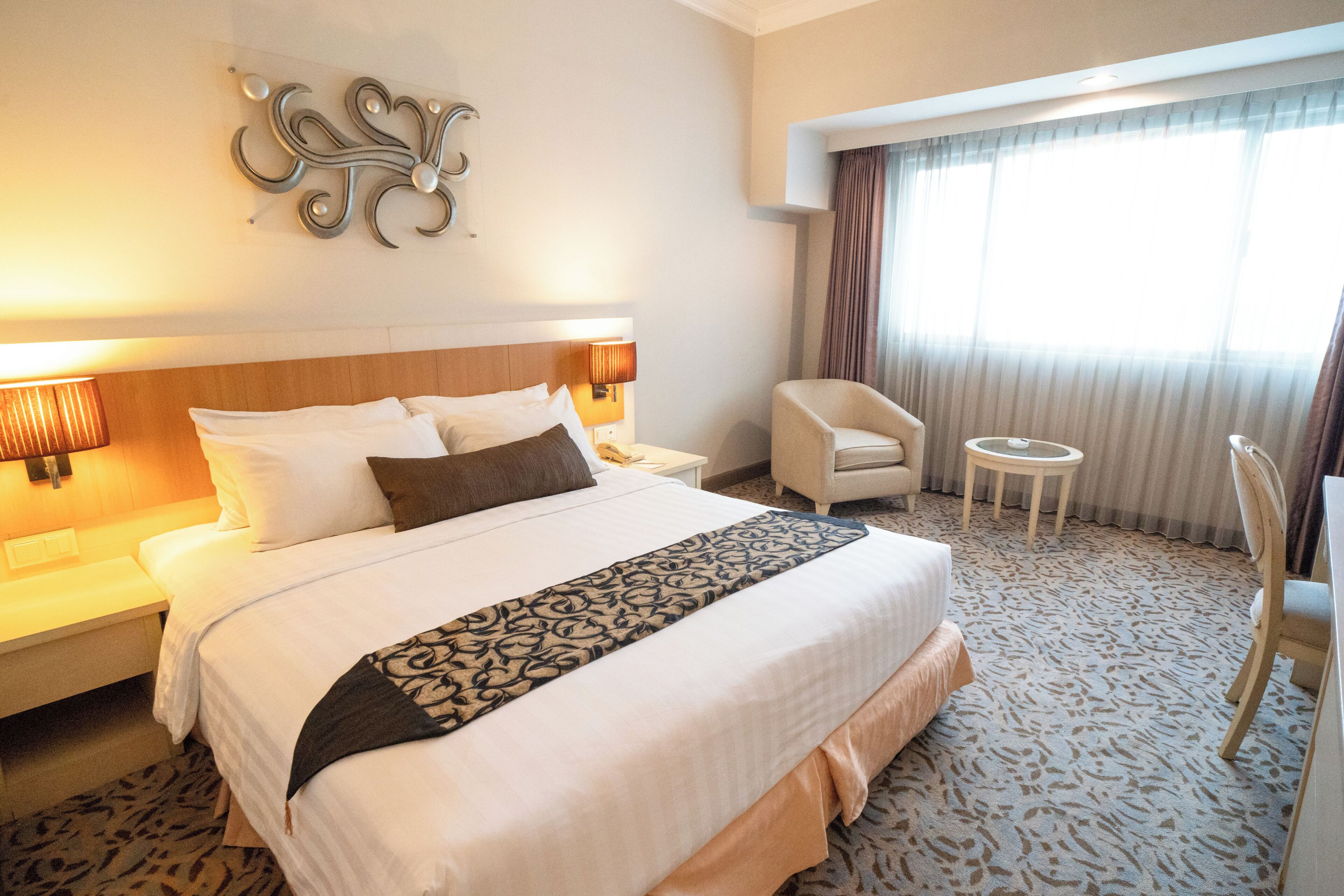 A hotel room with a large bed, a chair, a small table, and a decorative wall piece.