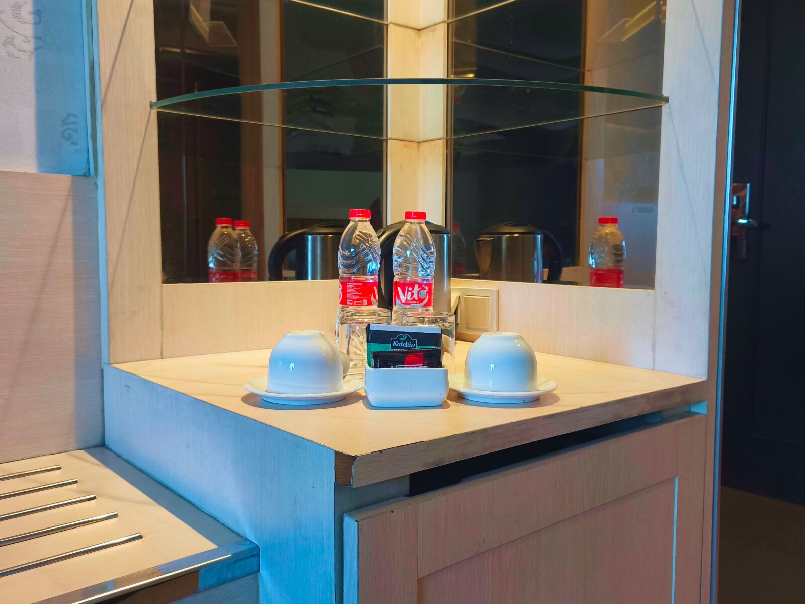 Une chambre d’hôtel avec un comptoir en bois, deux tasses blanches, un verre, une bouteille d’eau, une bouteille de Coca-Cola, une boîte de mouchoirs Kleenex et une machine à café.