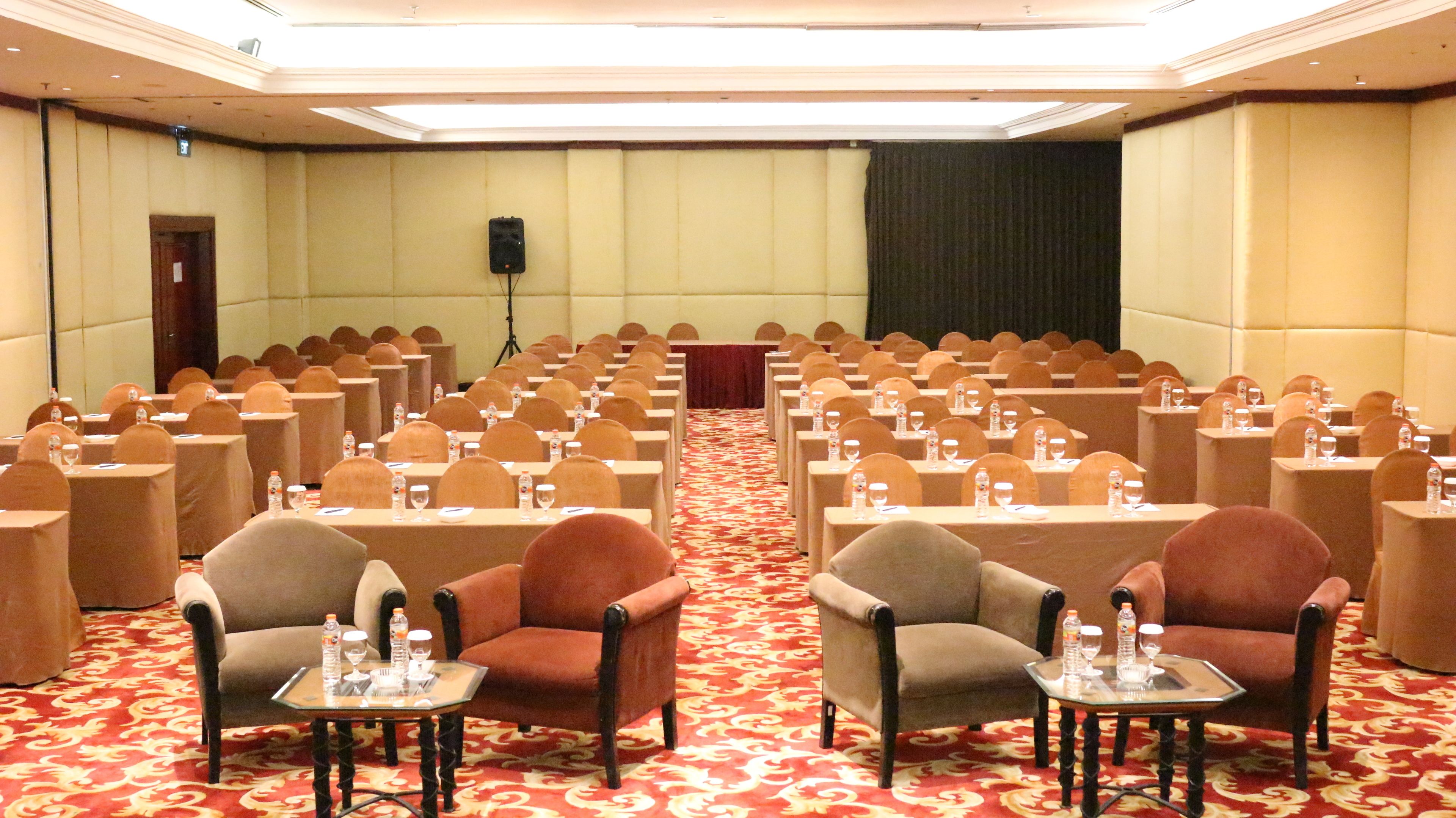 Un salón de conferencias con filas de sillas, mesas con botellas de agua y un escenario al frente.
