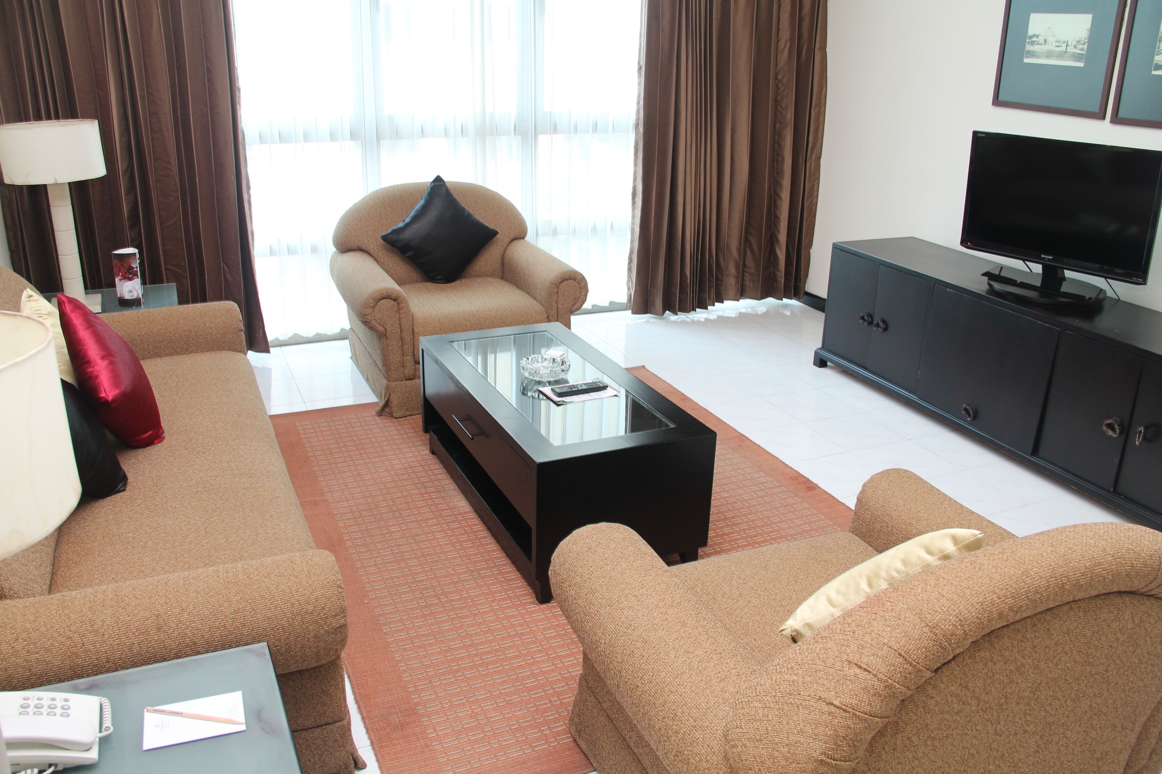 Habitación de hotel moderna con sofá color beige, una mesita de centro de cristal, televisor sobre un mueble y una ventana con cortinas.