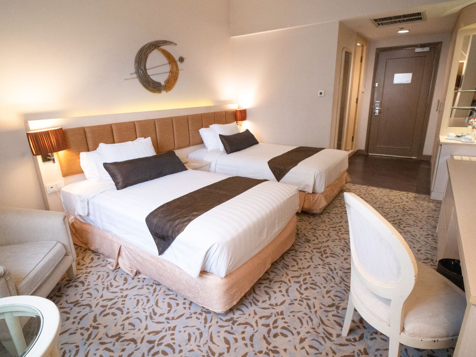 A hotel room with two beds, a headboard, bedside lamps, a patterned carpet, a white chair, and a door.
