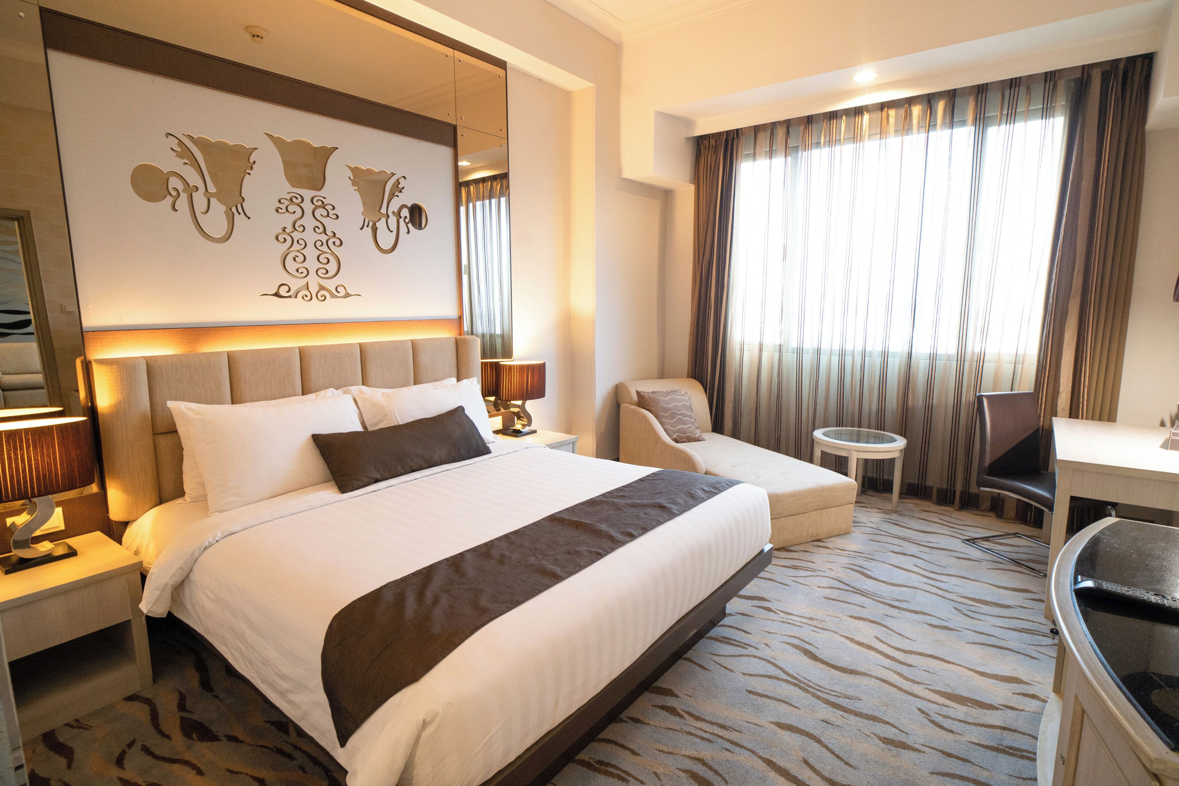 Habitación de hotel con una cama grande, un escritorio, una silla, una mesita pequeña y un ventanal con cortinas.