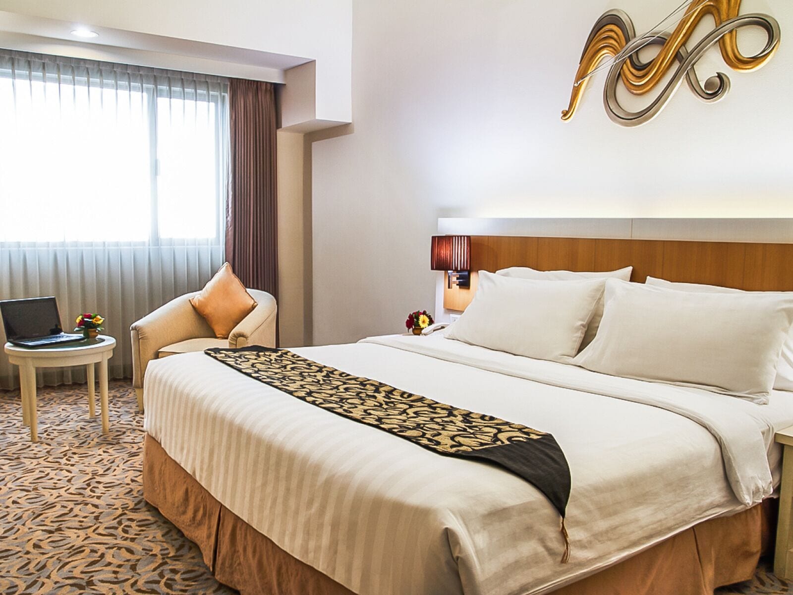 A hotel room with a large bed, a bedside table with a lamp, a laptop, and a view of the outside through a window with curtains.