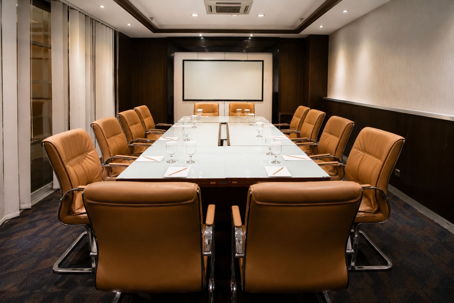 A conference room with a long table, brown leather chairs, a projector screen, and a whiteboard.