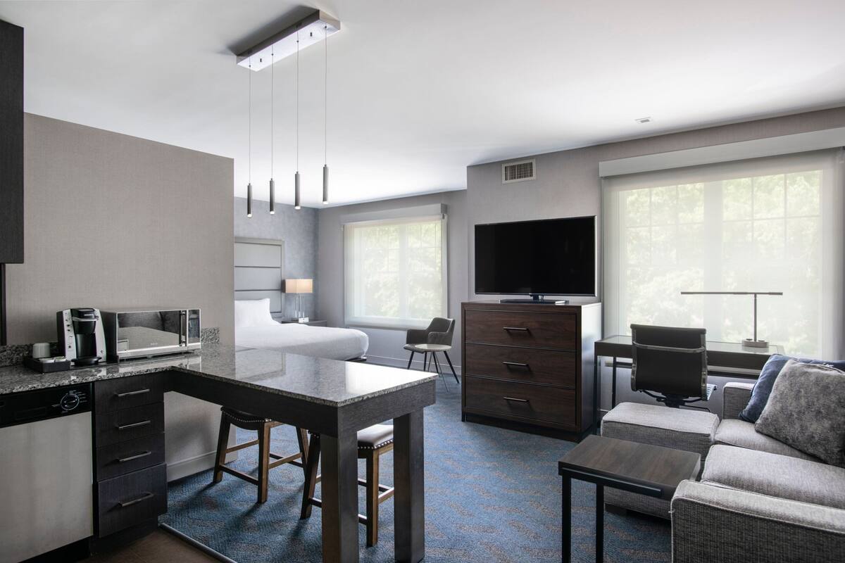 A modern hotel room with a kitchen area, a dining table, a sofa, and a TV.