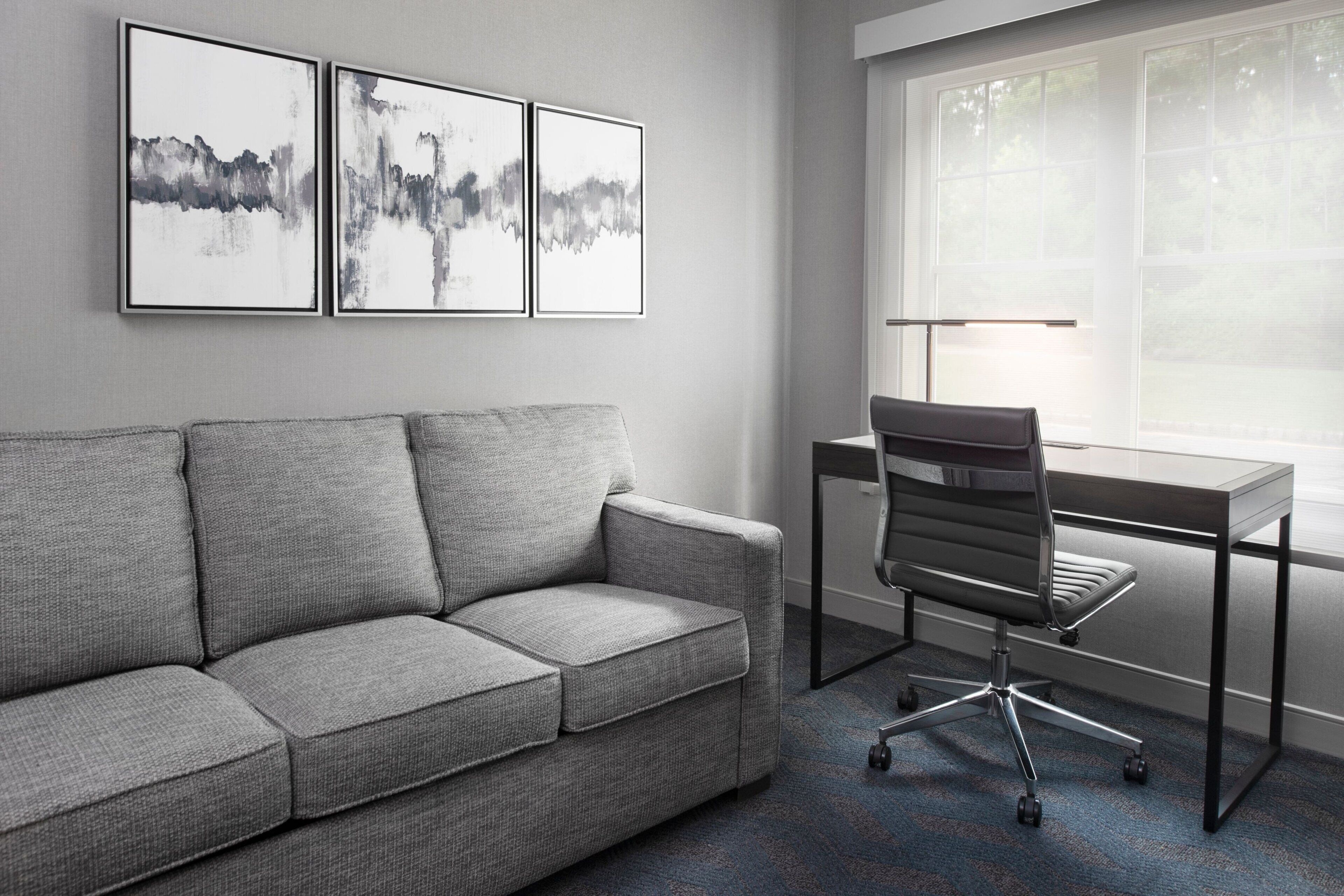 A modern office space with a grey sofa, a desk with a chair, and three abstract paintings on the wall.