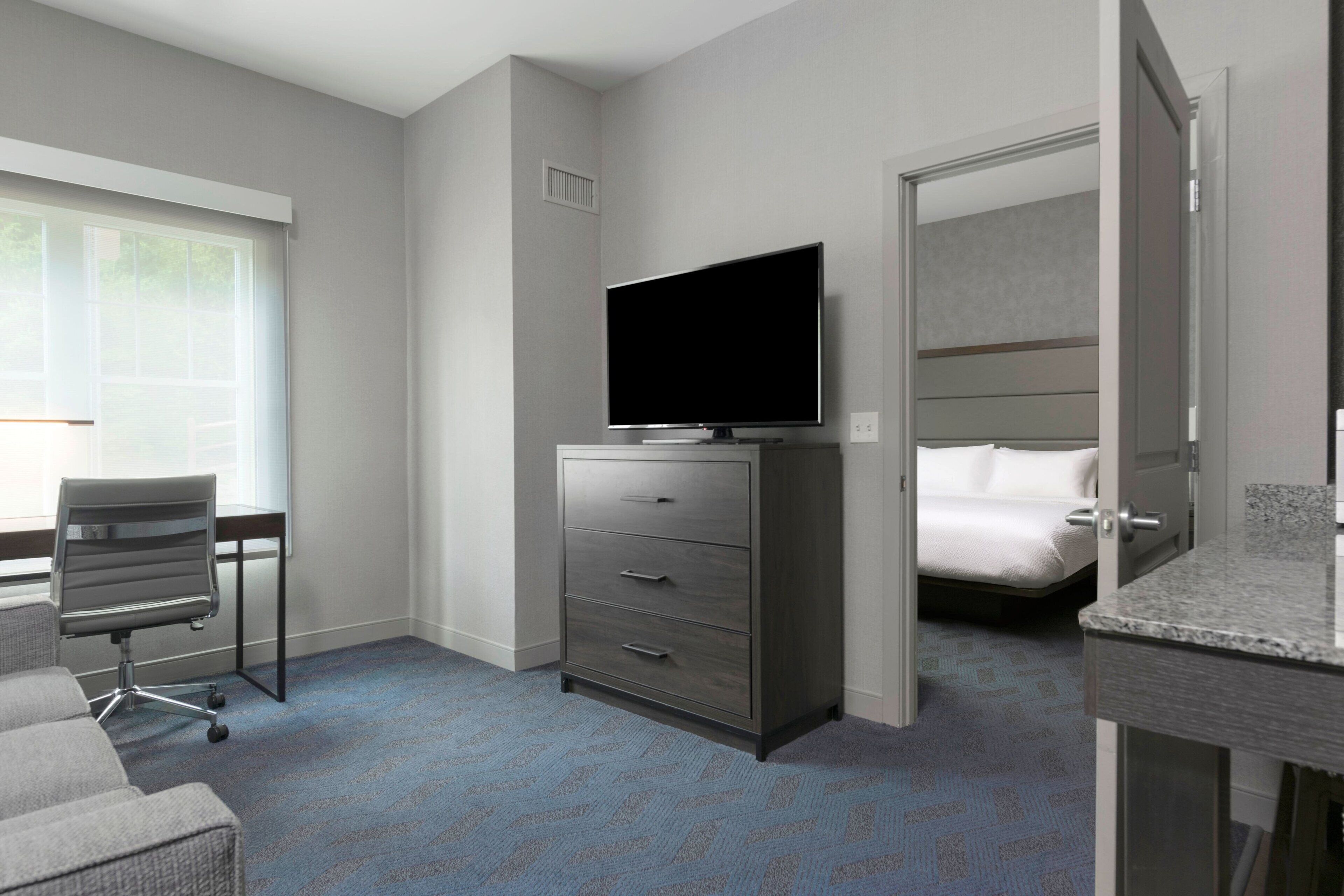 Quarto de hotel com uma cama, televisão em uma cômoda, uma escrivaninha e uma cadeira.