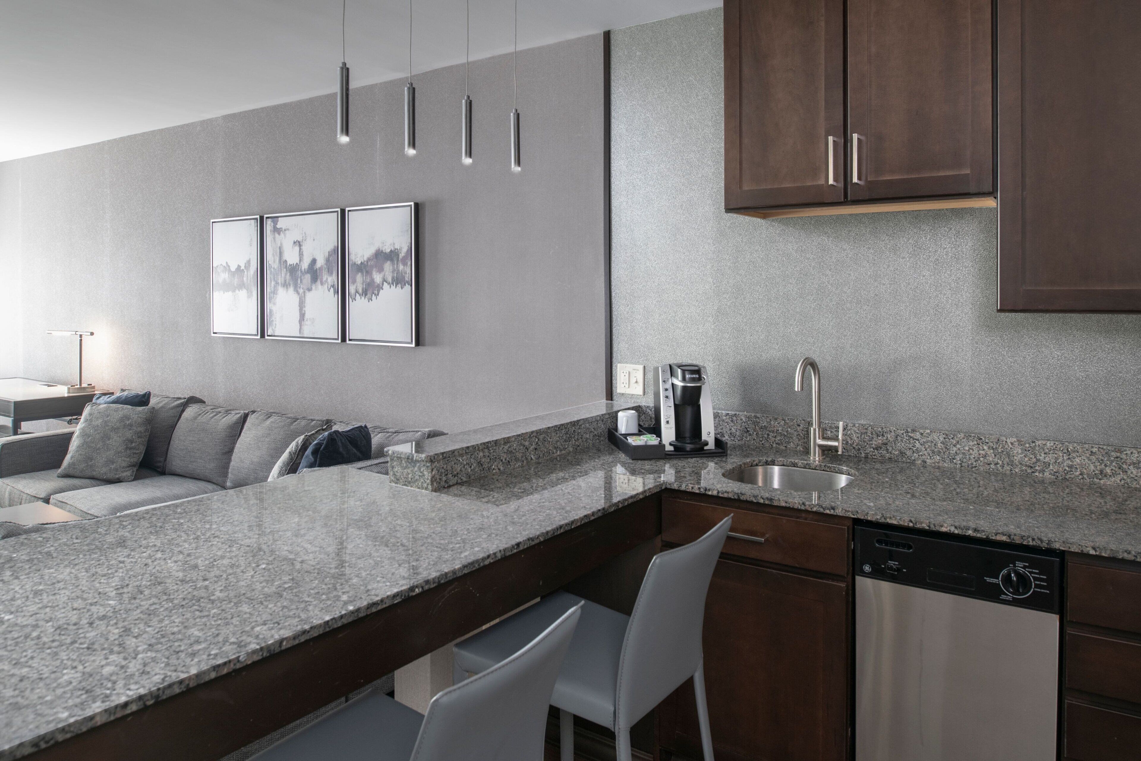 A modern kitchen with a granite countertop, stainless steel appliances, and a dishwasher. There is a bar area with white chairs and a coffee machine.