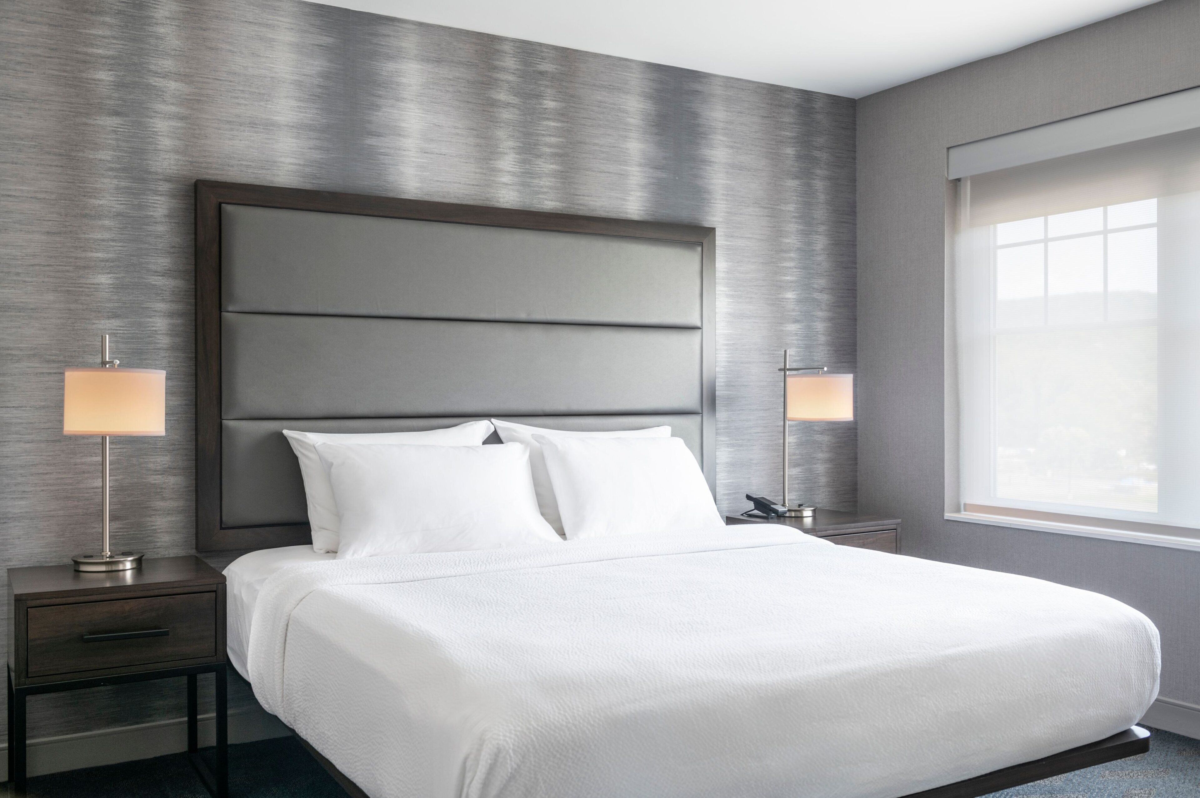 A modern hotel room with a large bed, two bedside tables with lamps, and a large window providing natural light.