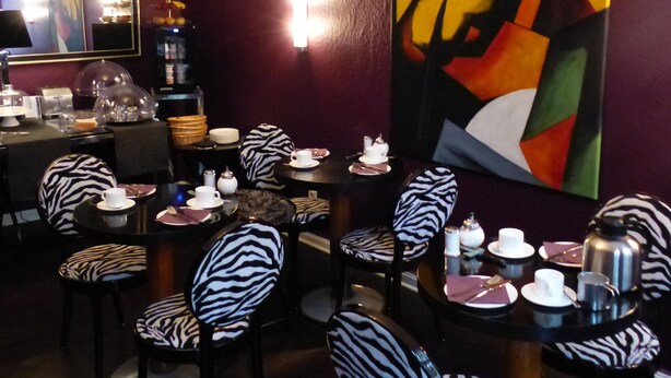 Un comedor con sillas a rayas de cebra, un cuadro abstracto grande en la pared y una mesa dispuesta con platos, tazas y una cafetera.