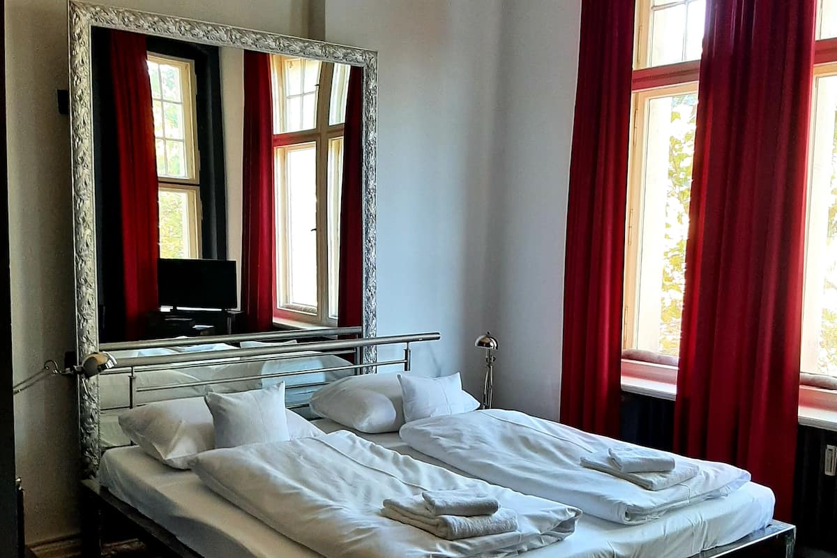 A bed with white linens, a large mirror above it, and red curtains on the windows.