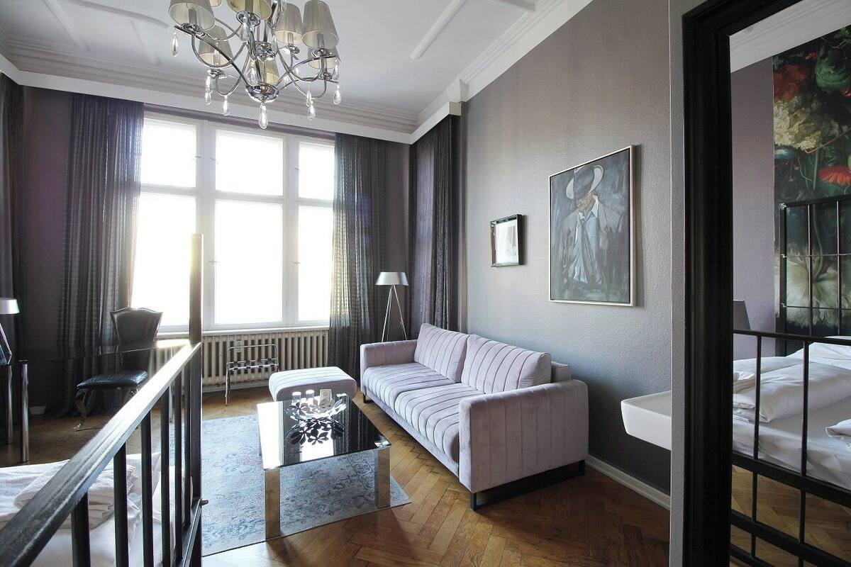 A modern living room with a sofa, a coffee table, and a chandelier. There is a window with curtains and a radiator.