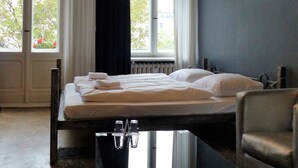Une chambre d’hôtel avec un lit, une chaise, un lustre, de grandes fenêtres et une vue sur les arbres.