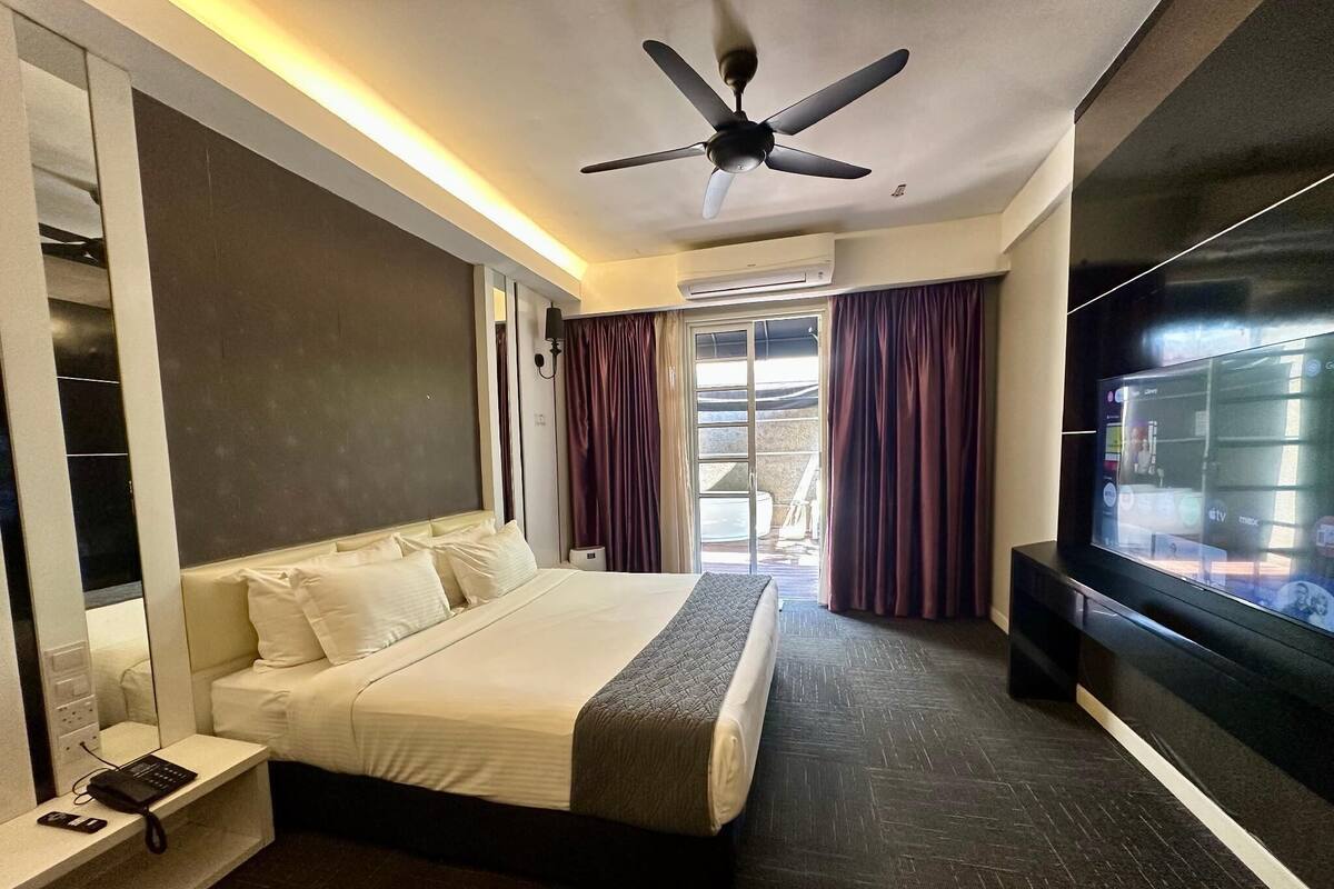 A modern hotel room with a large bed, a flat-screen TV, a ceiling fan, and a view of the outdoors through a sliding door.
