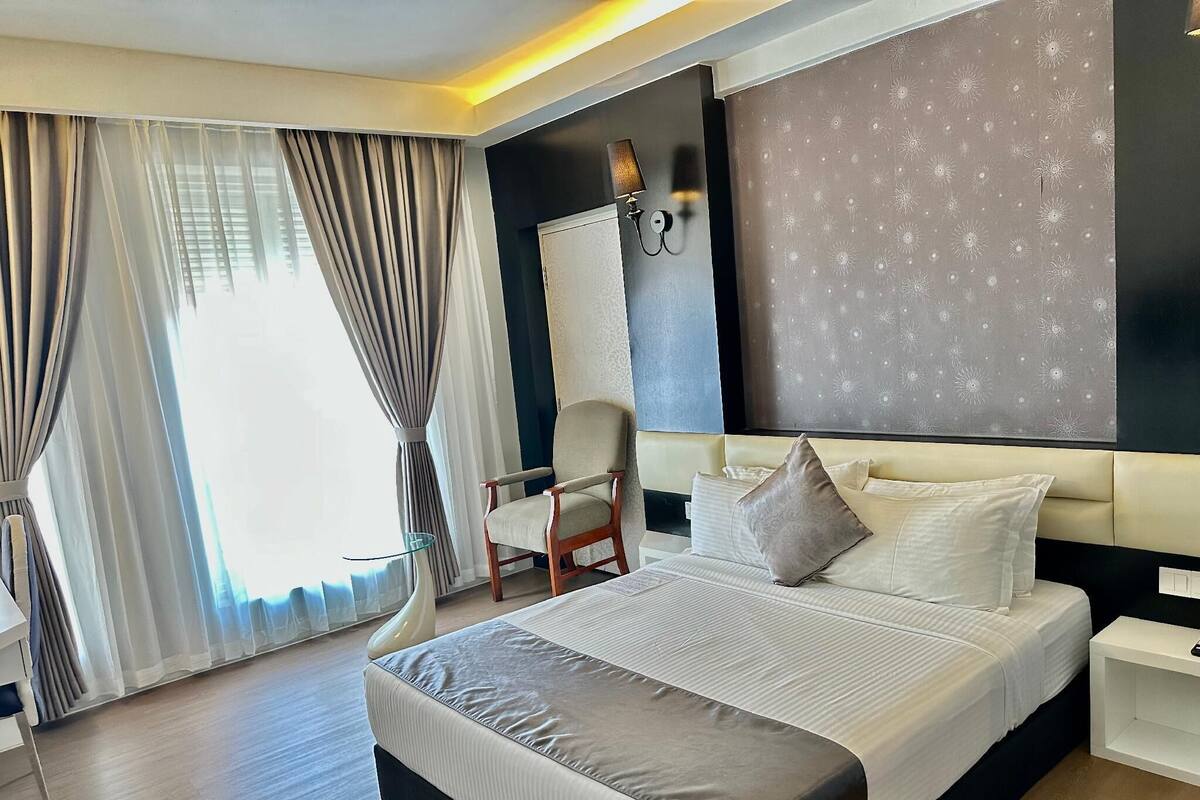 A modern hotel room with a large bed, a chair, a nightstand, and a wall-mounted lamp.