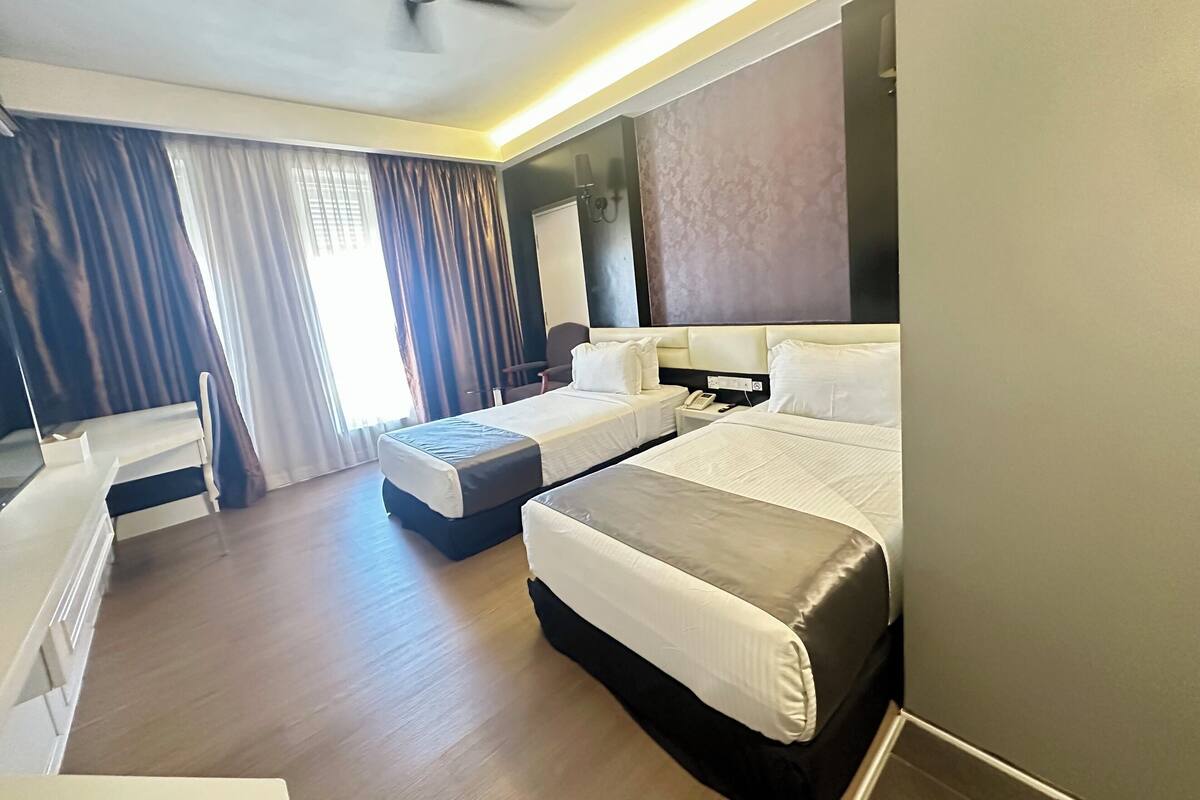 A hotel room with two beds, a desk, a chair, a ceiling fan, and a large window with curtains.