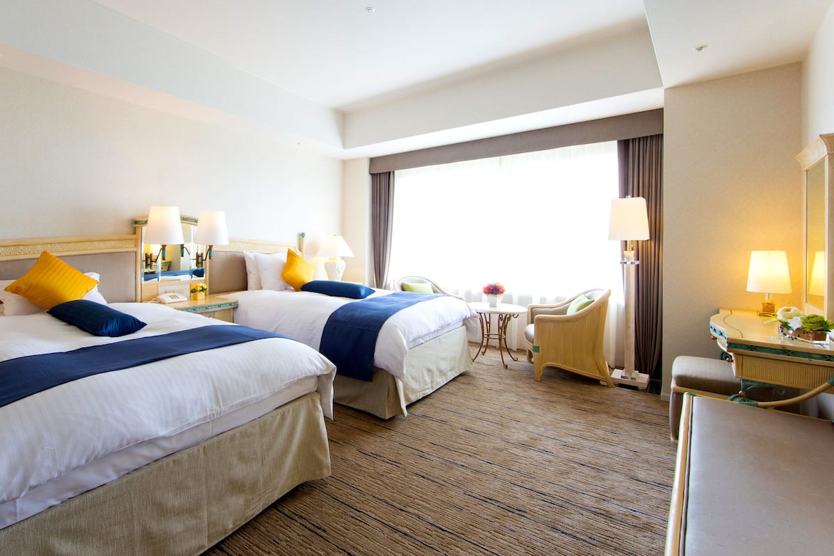 A hotel room with two beds, a small table, a chair, and a desk with a lamp.