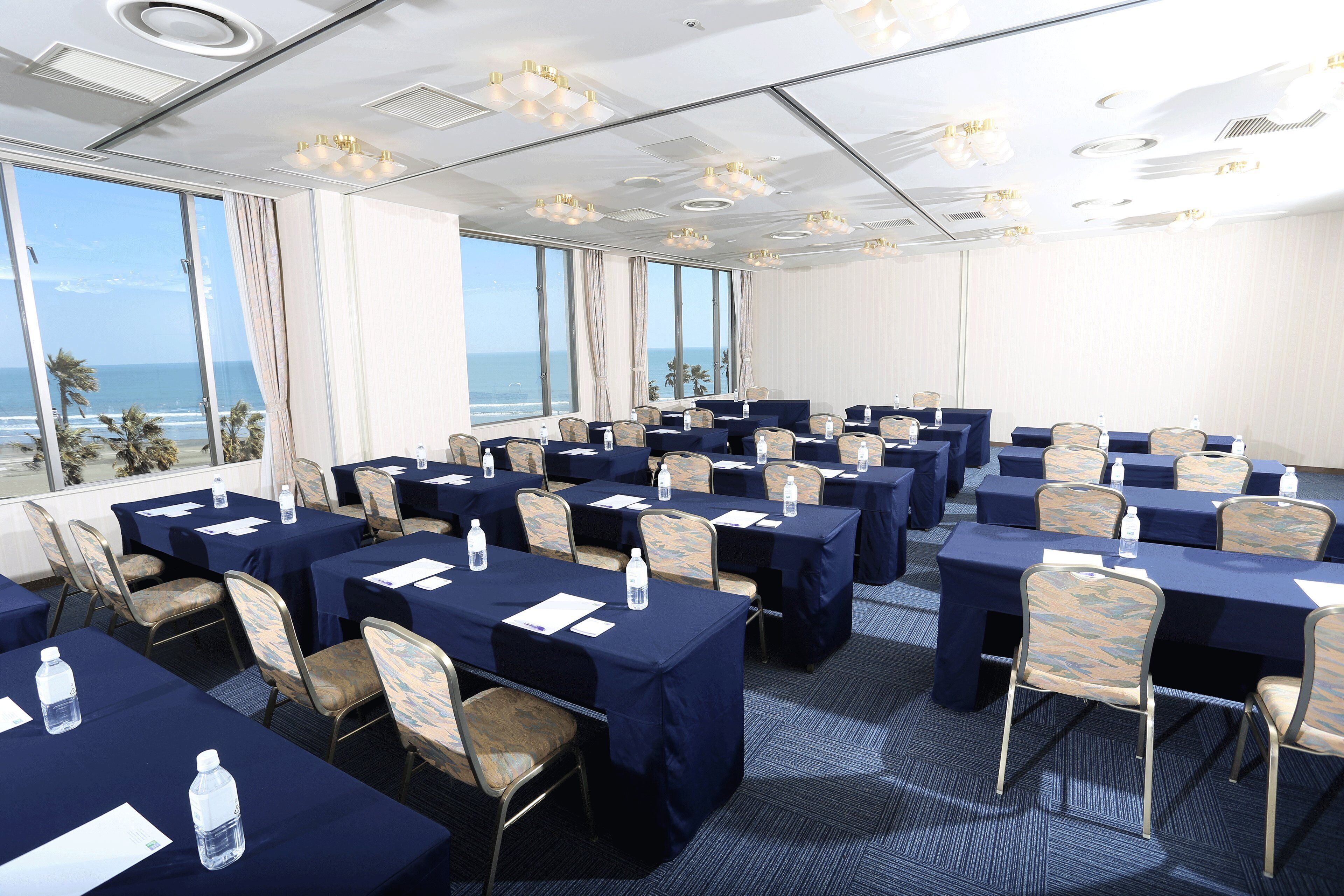 Een congreszaal met tafels en stoelen, grote ramen met uitzicht op de oceaan en plafondlampen.