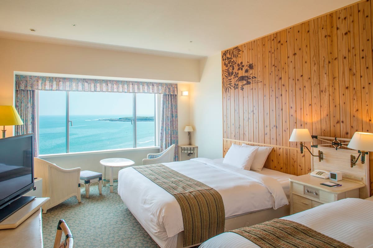 A hotel room with two beds, a TV, a small table, and a window with a view of the sea.