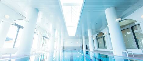 A swimming pool with clear blue tiles, surrounded by white columns and large windows allowing natural light.