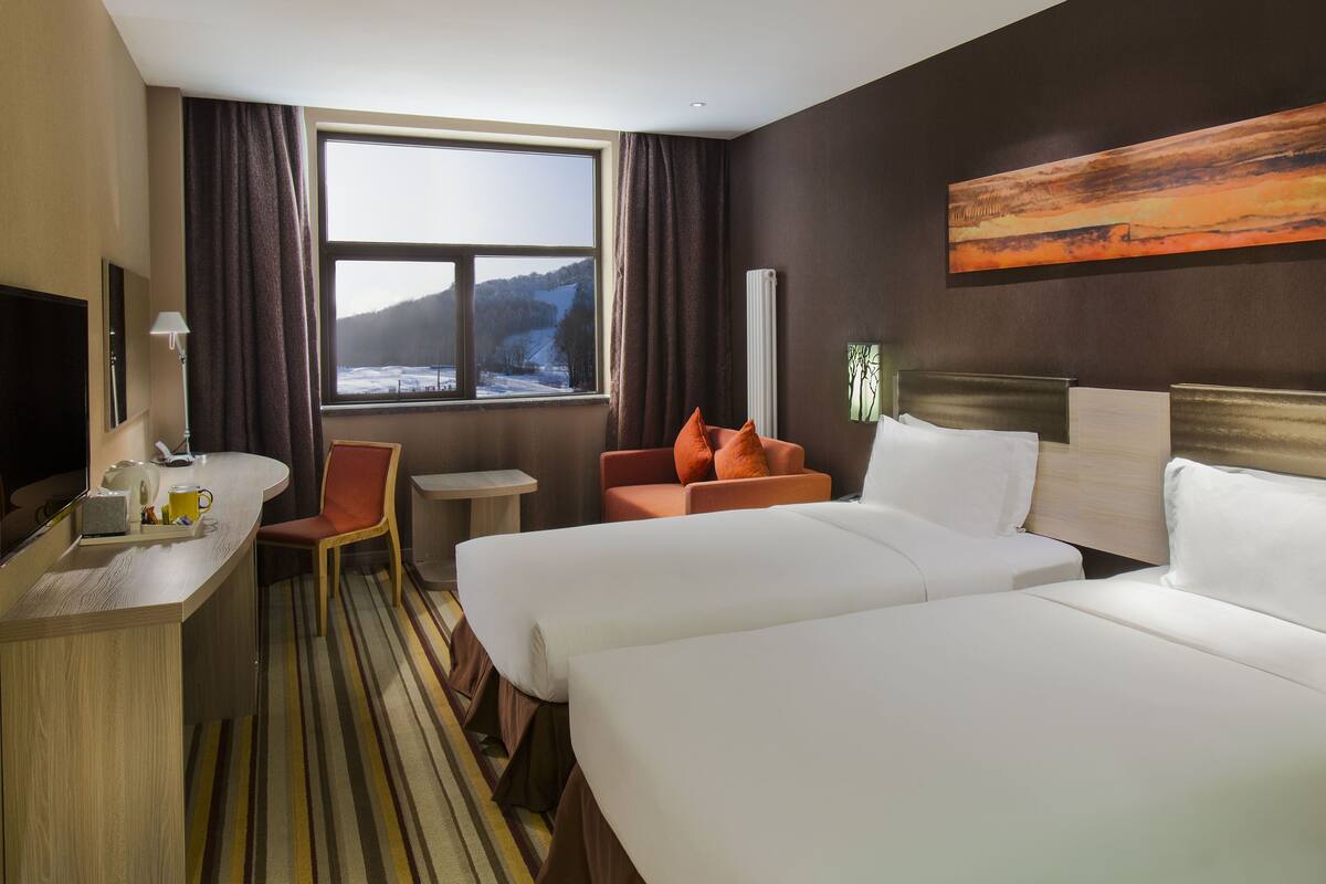 A hotel room with two beds, a desk, a chair, a window with a view of a snowy landscape, and a painting on the wall.
