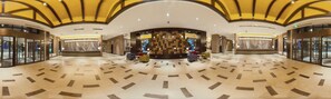 A spacious lobby with a geometric floor pattern, a central seating area with colorful chairs, and a decorative wall with a tree sculpture.