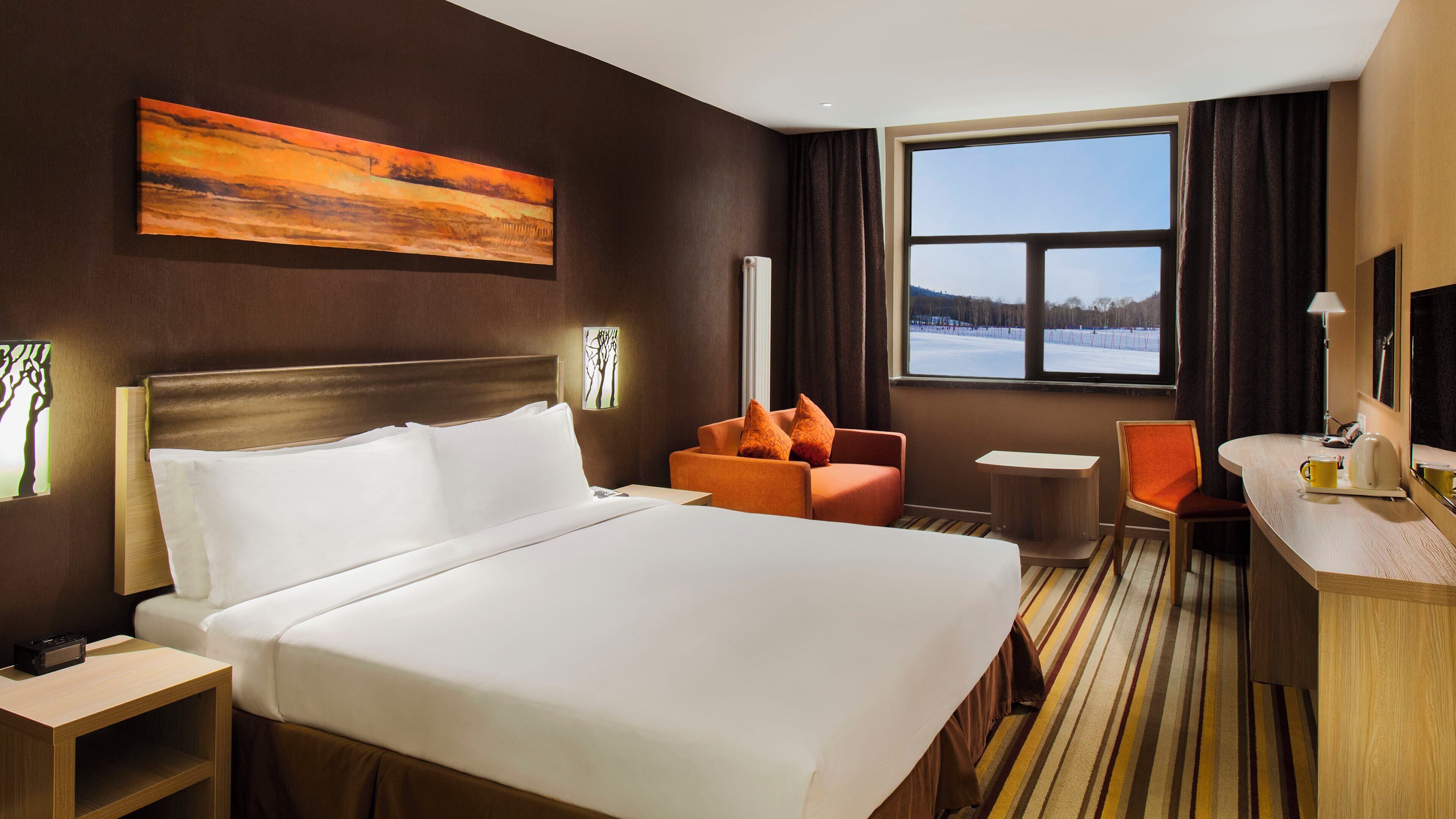 Habitación de hotel con una cama grande, un escritorio, una silla y un ventanal con vistas a un paisaje nevado.